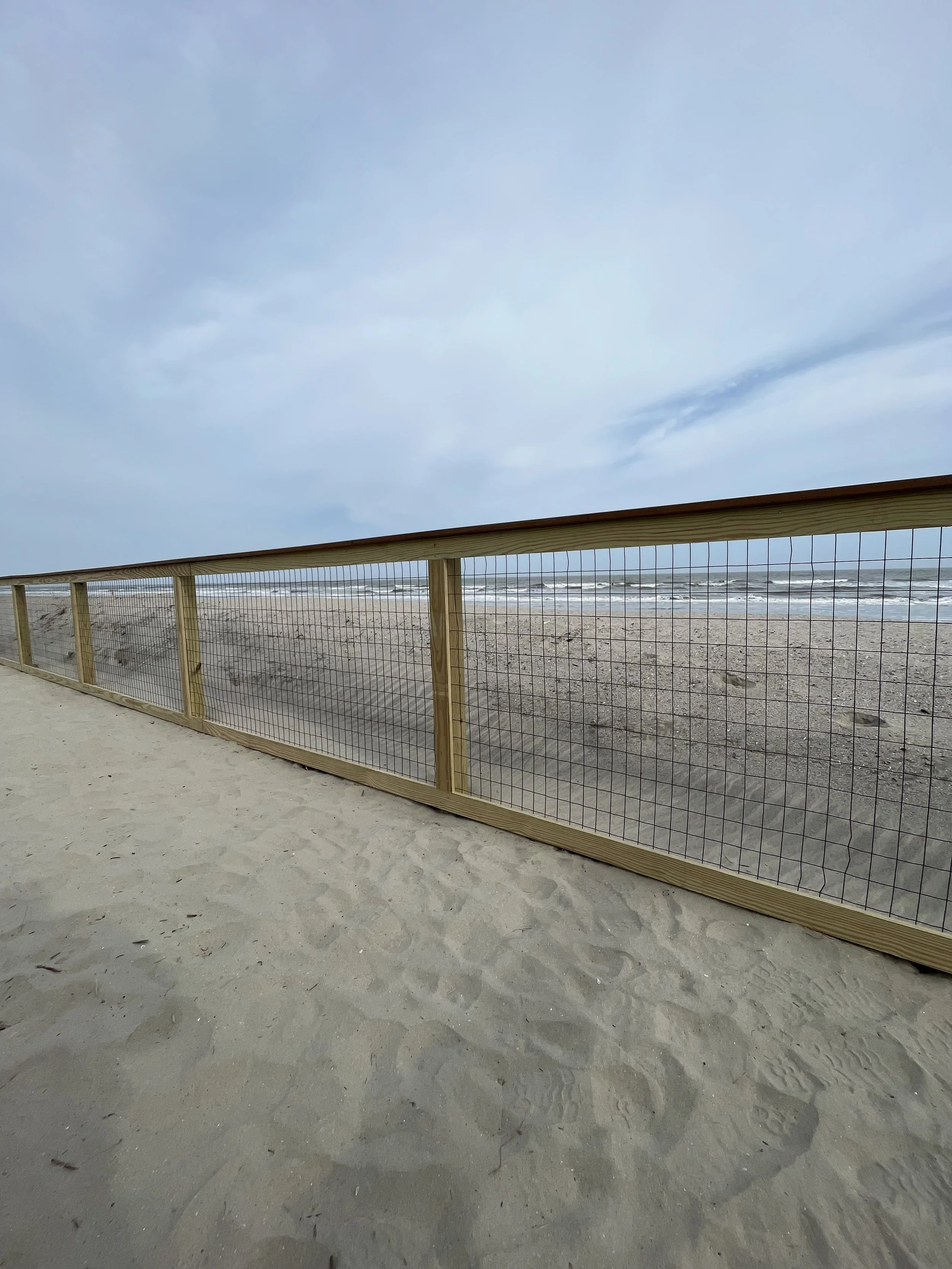 Wooden and wire living fence along a sandy beach with ocean waves and a cloudy sky in the background.
