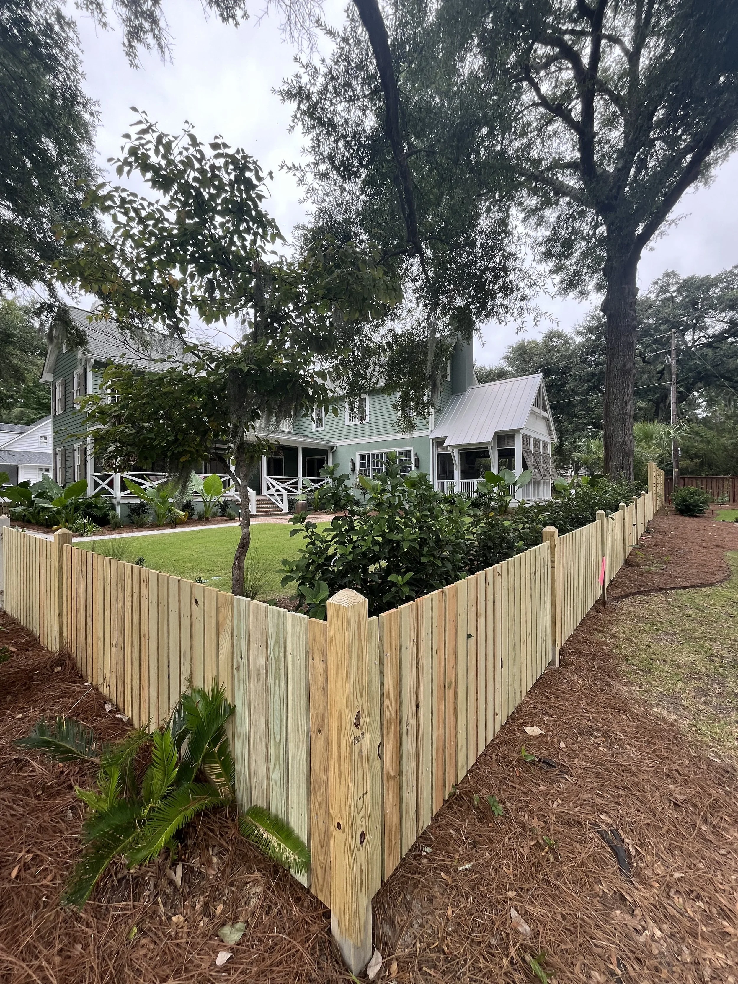 A new wooden fence with vertical planks surrounds a lush green yard with trees and shrubs in front of a light-colored house with a porch and bay window, under an overcast sky.