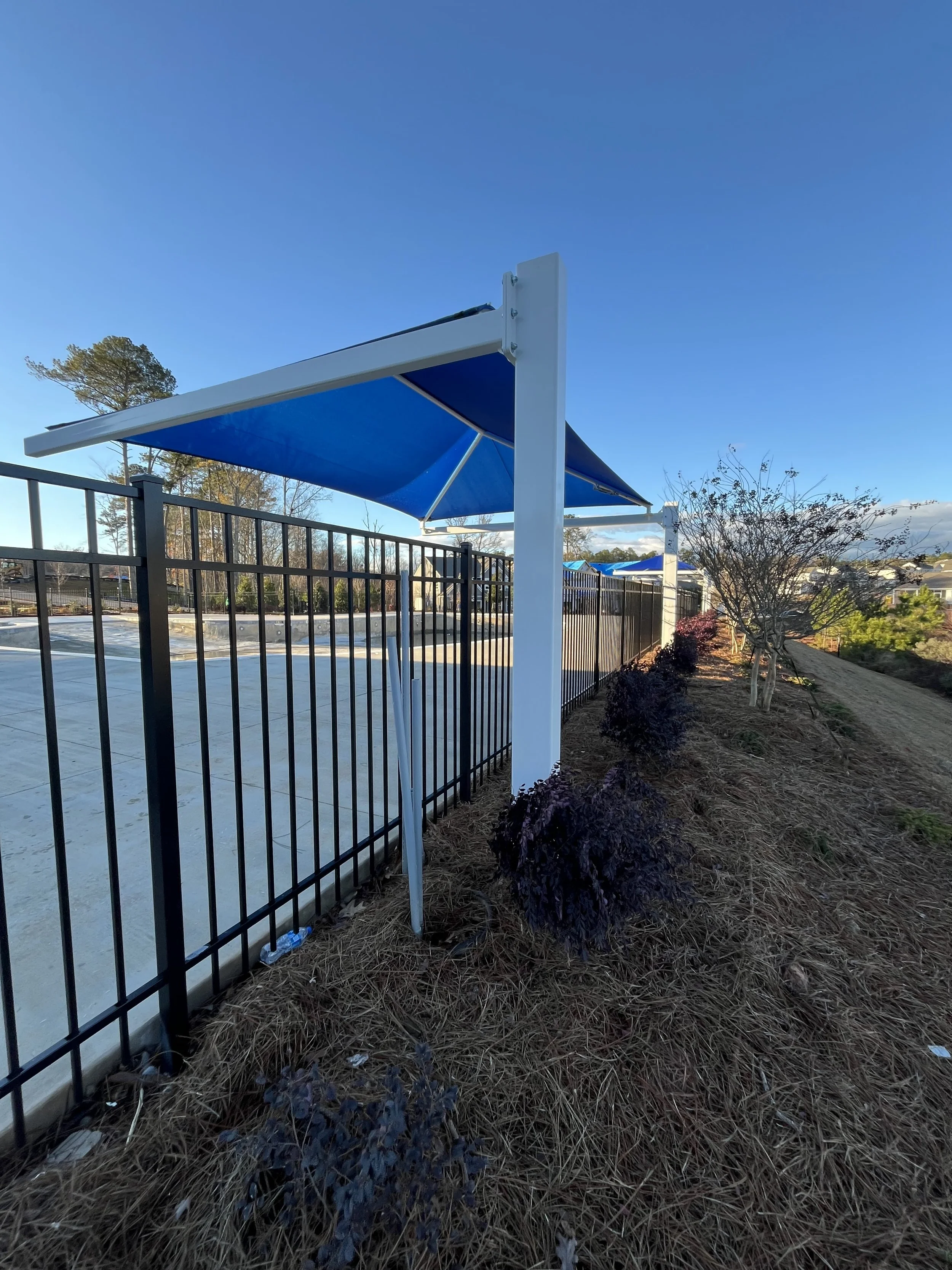 A black aluminum fence running along a sidewalk with a blue canopy above it. There are trees and shrubs nearby, with a clear blue sky.