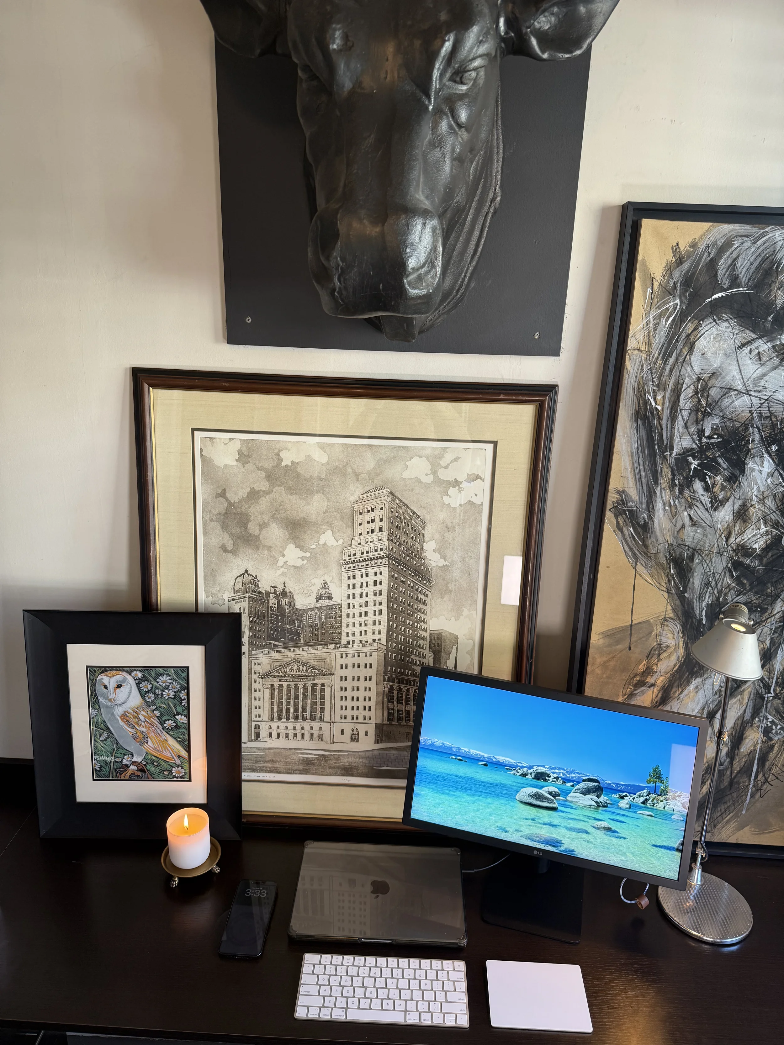 A workspace with various framed artwork, a computer monitor displaying a beach scene, a candle, an iPad, a phone, a keyboard, a mouse, and a lamp on a dark desk.