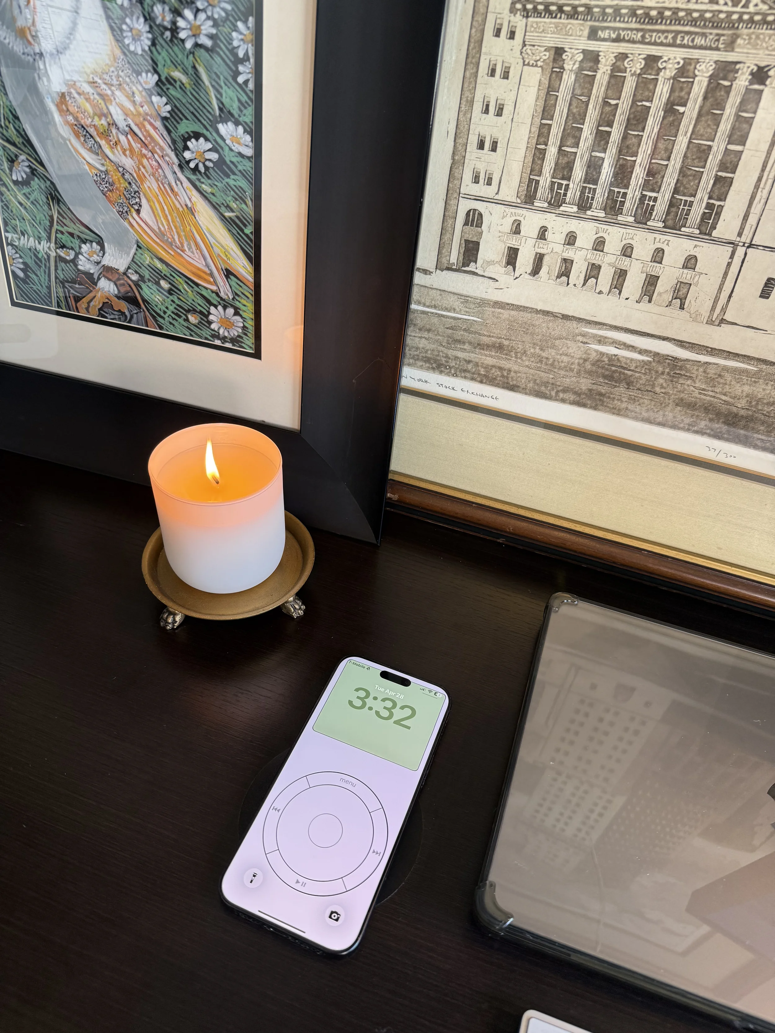 A black table with a lit candle in a brass holder, a smartphone displaying a remote control app showing the time 3:32, and framed artwork of a building and a bird.