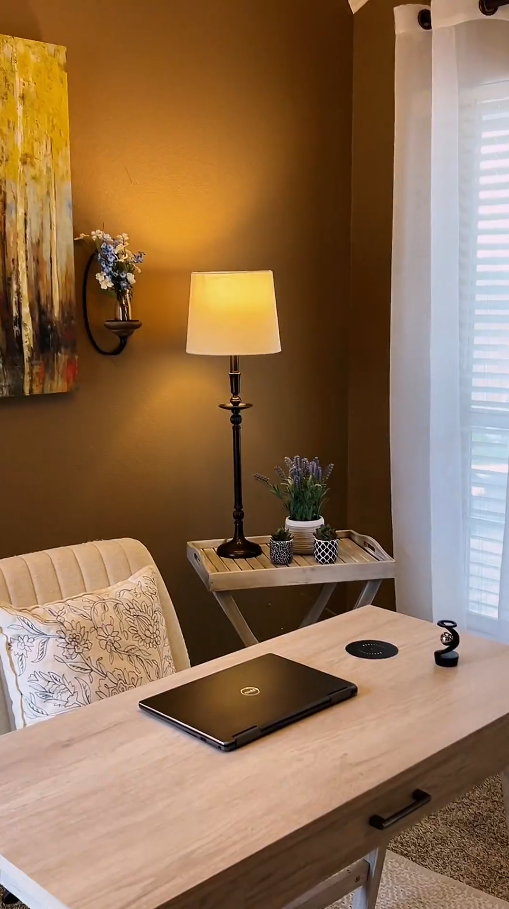A cozy corner of a room with a wooden table, a laptop, a beige chair with floral pillow, a side table with a lamp, potted plants, and a wall-mounted shelf with flowers, near a window with white curtains.
