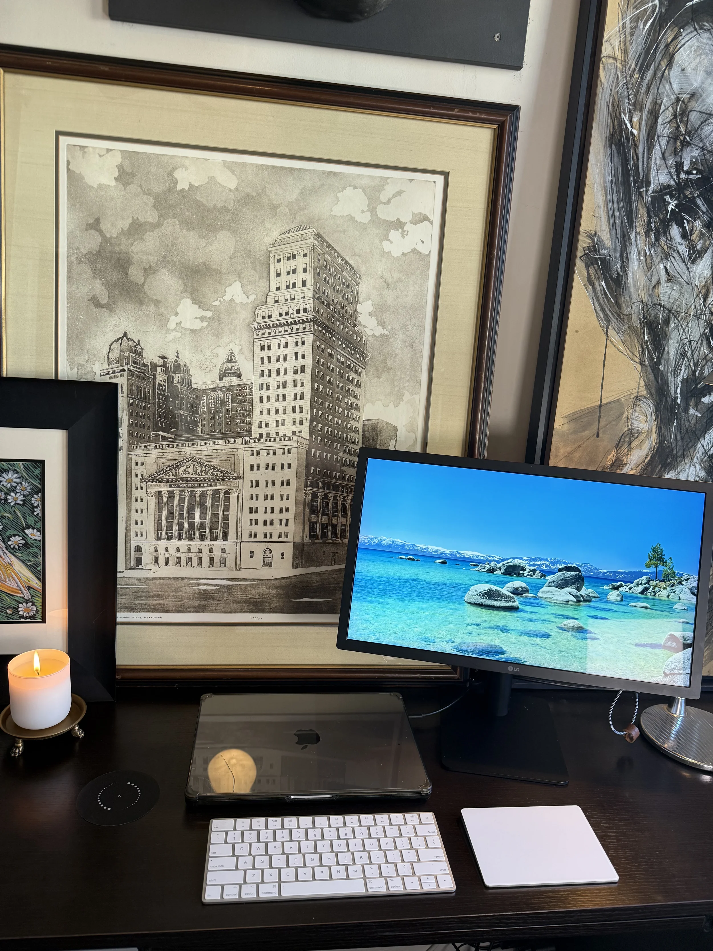 A home office desk setup including a computer monitor displaying a beach scene with rocks and mountains, an Apple Magic Keyboard, a Trackpad, a MacBook, a candle, and framed artwork of a cityscape with tall buildings, clouds, and architectural details.