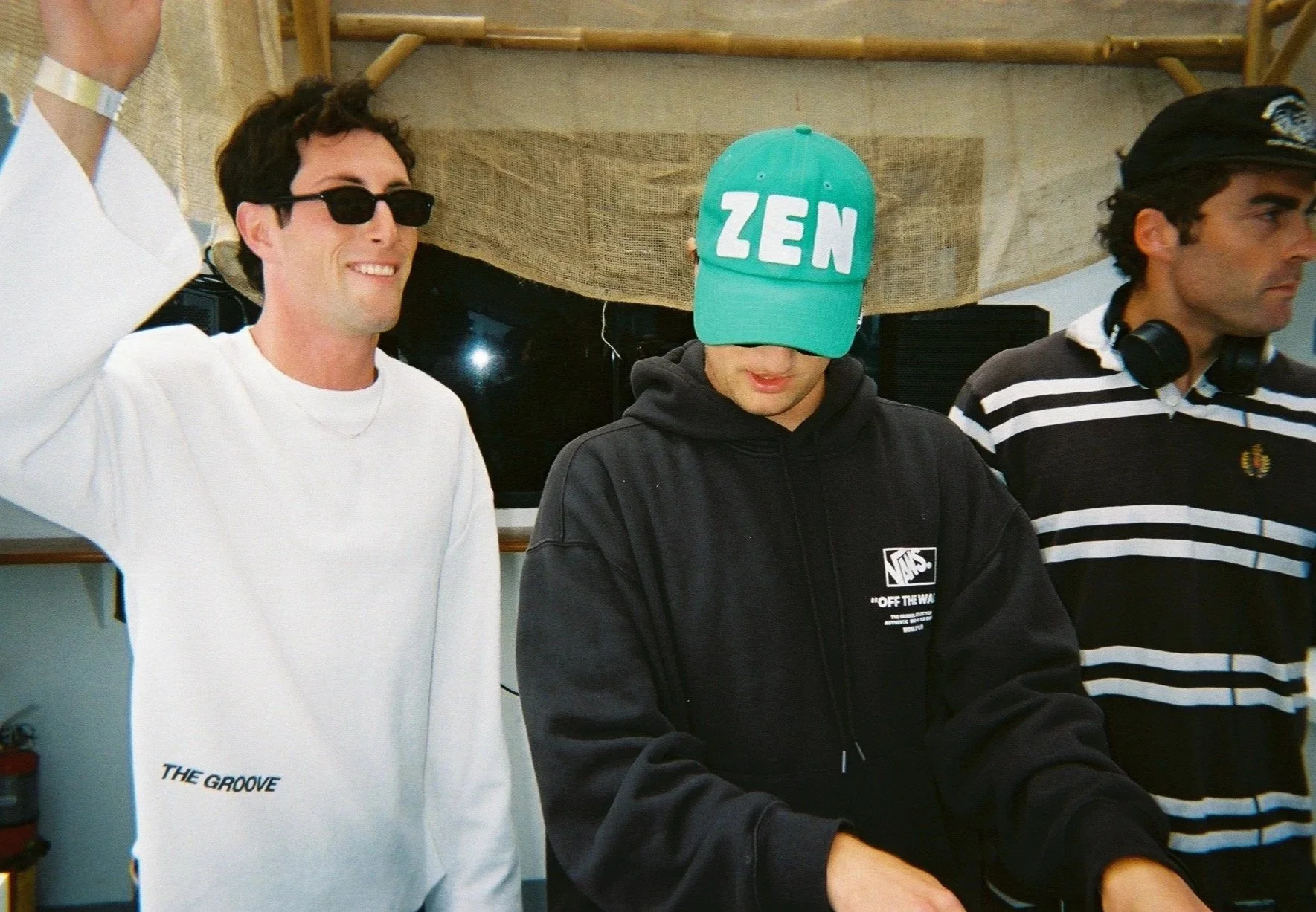 Three young men standing together. The man on the left is smiling, wearing black sunglasses and a white sweatshirt. The man in the middle is looking down, wearing a lime green cap with the word "ZEN" on it and a black hoodie. The man on the right is looking to the side, wearing a black + white striped polo shirt and headphones around his neck.