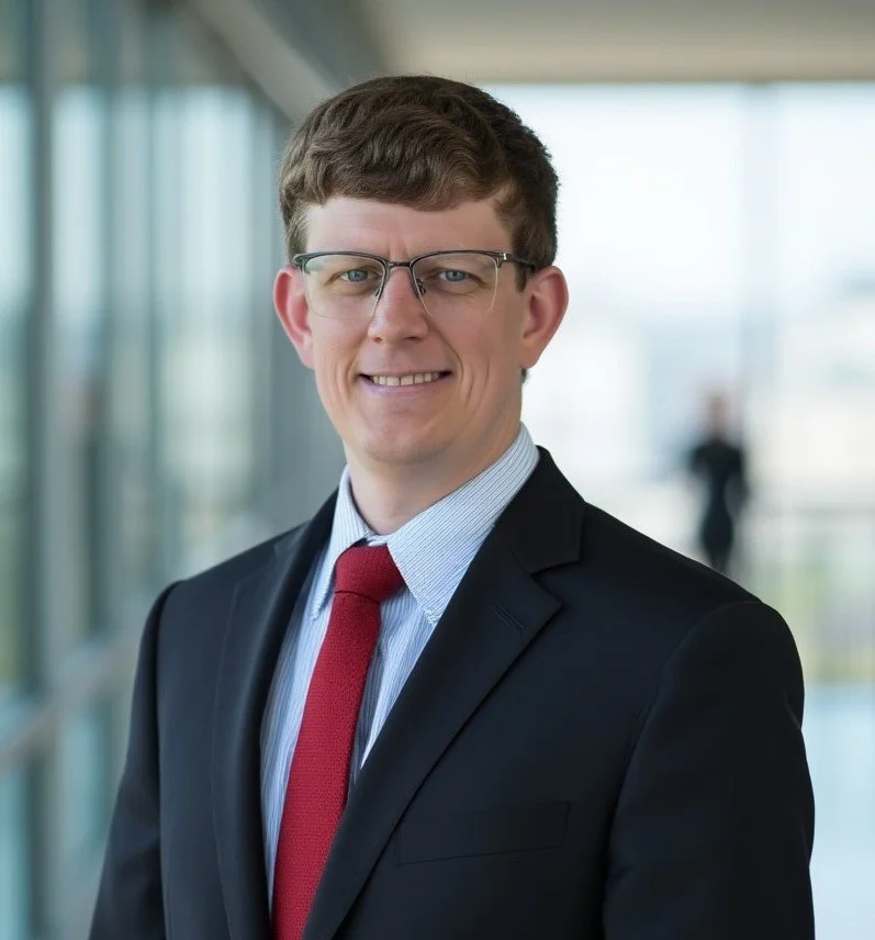 A man in a business suit wearing glasses, smiling, indoors near large windows.