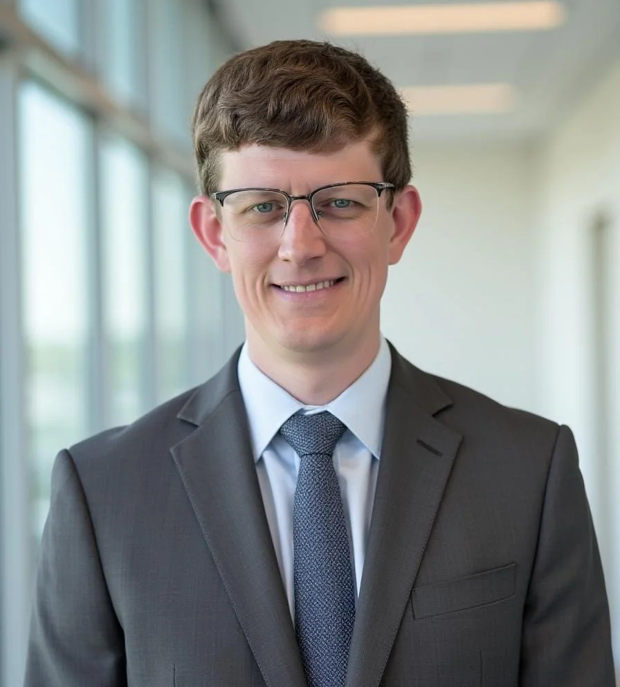 A young man wearing glasses, a dark suit, a light blue shirt, and a patterned tie standing in a modern, well-lit office corridor with large windows.