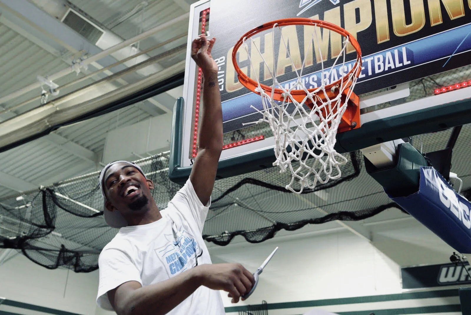 A joyful basketball player wearing a white t-shirt and a backward cap celebrates under a basketball hoop inside a gymnasium, holding a trophy in his right hand.