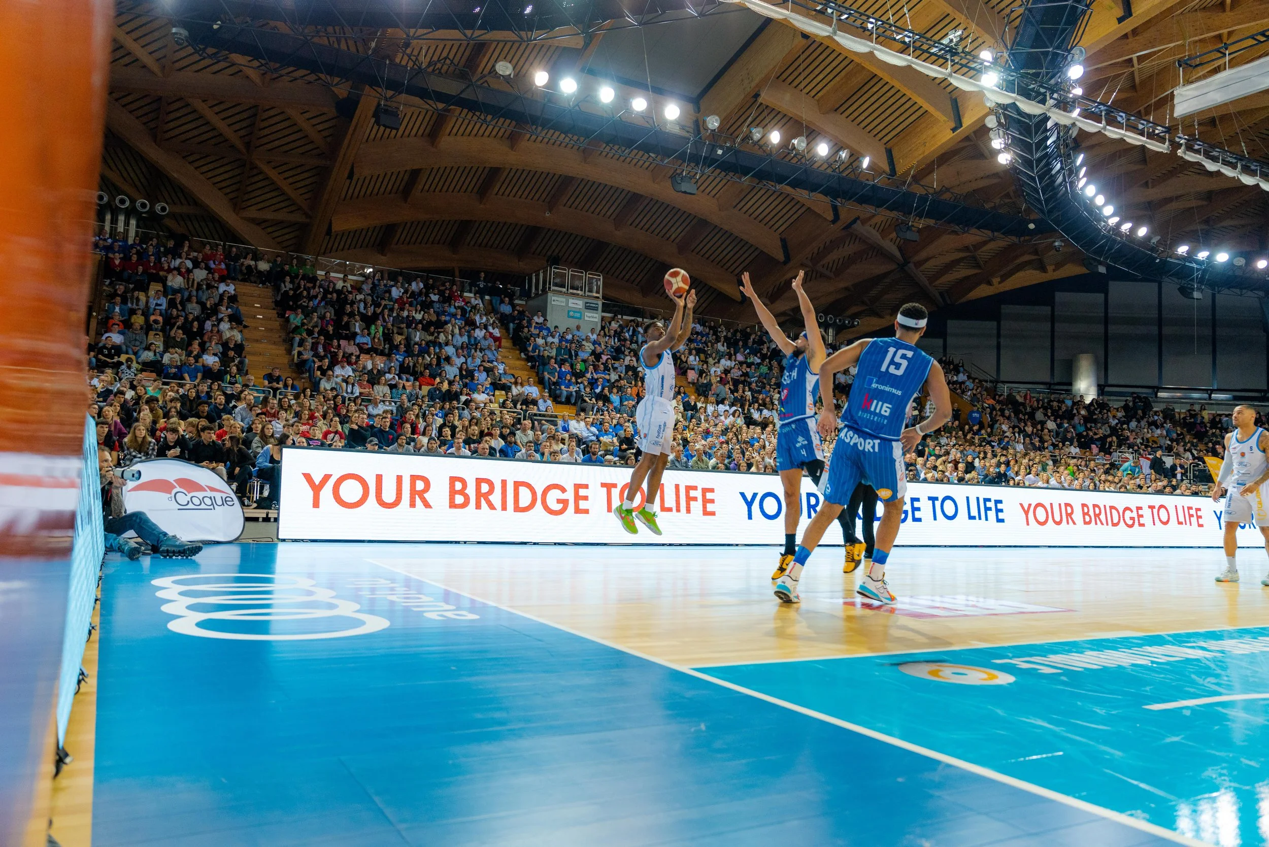 A basketball game in progress with players in action on a court filled with spectators and advertising banners in the background.