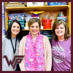 Three smiling women standing in front of a colorful classroom shelf. The woman on the left has dark hair, the woman in the middle has light hair and is wearing a pink scarf, and the woman on the right has light hair and is wearing a purple top. There is a school logo in the bottom left corner.