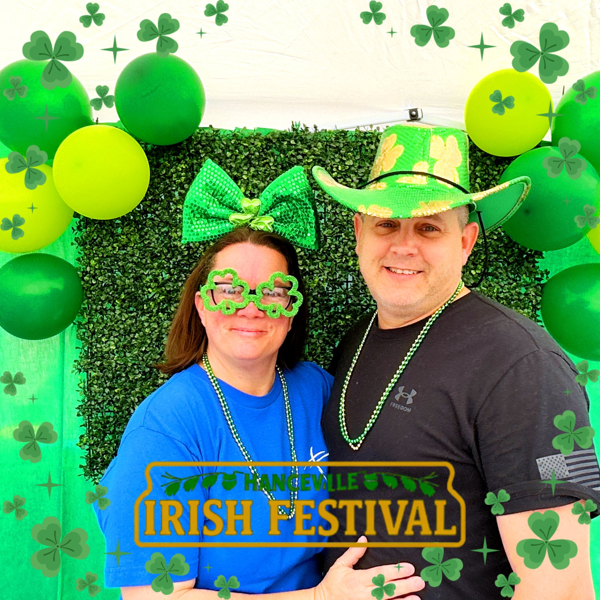 A woman and a man celebrating St. Patrick's Day at a festival, both wearing green accessories like shamrock glasses and a large bow and hat. They are standing in front of a festive backdrop decorated with green balloons, shamrocks, and greenery, with a sign that reads 'HANCEVILLE IRISH FESTIVAL'.