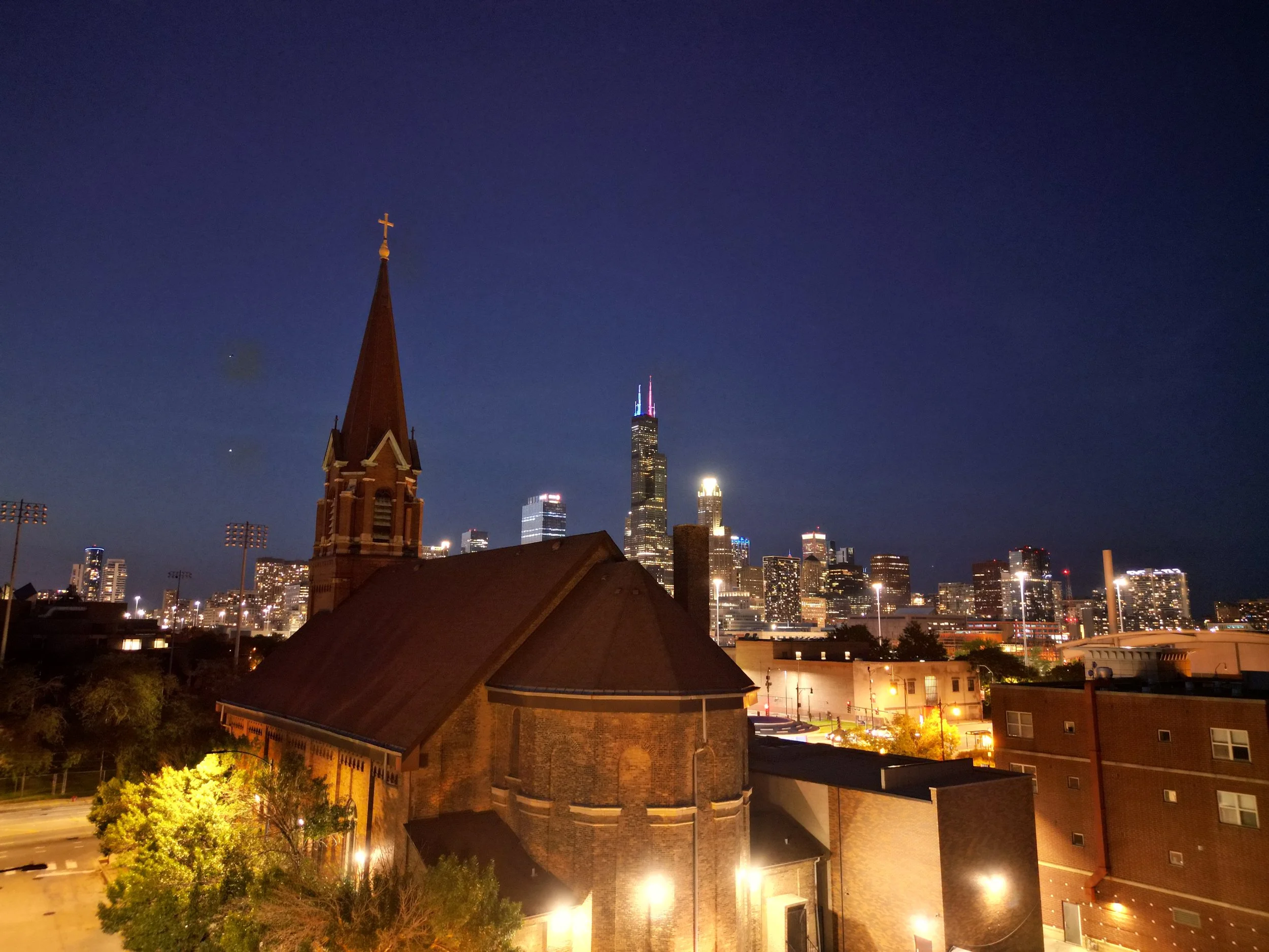 Vista nocturna del skyline de un centro urbano con edificios altos iluminados y un cielo oscuro, se destaca una iglesia con aguja y cruz en primer plano, y en el fondo, la Torre Willis con luces rosas y azules.