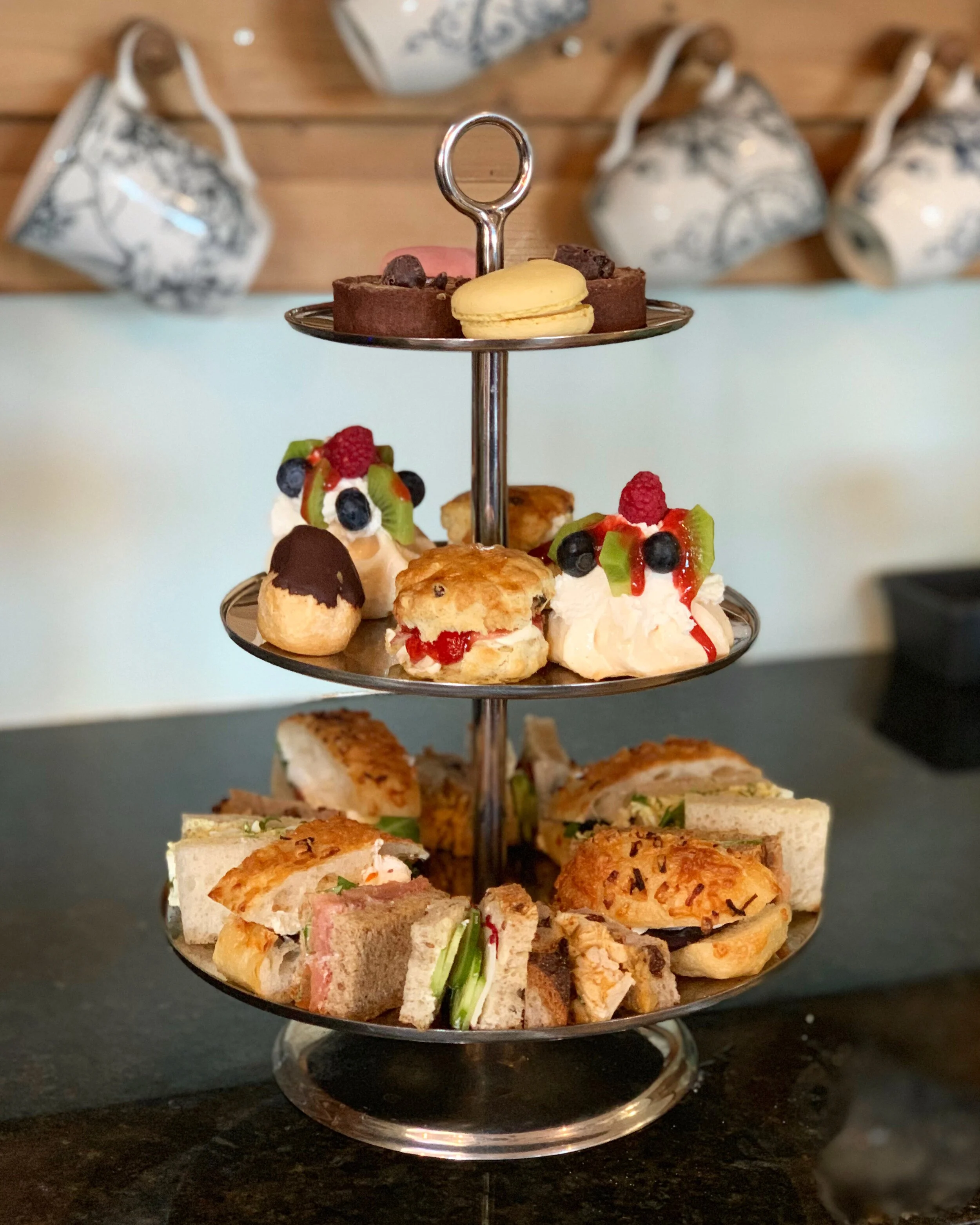 Three-tiered tray with assorted desserts and finger sandwiches on a black countertop, with several decorative coffee mugs hanging on the wall in the background.