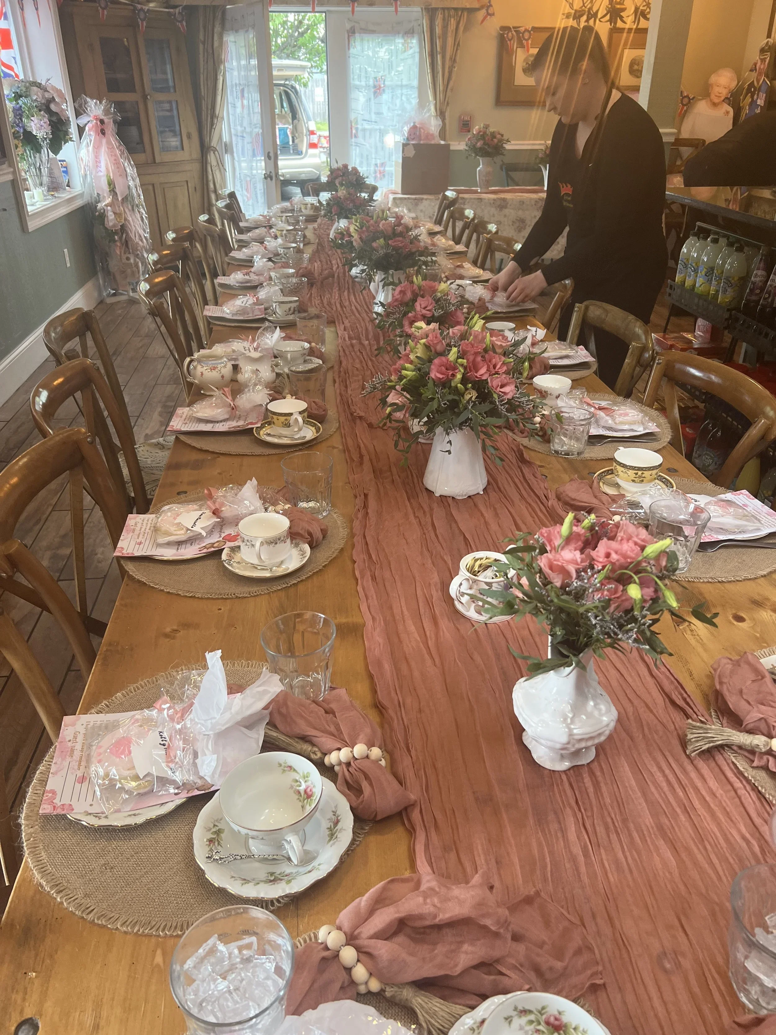 A long dining table set for a celebration with floral centerpieces, tea cups, glasses, and decorations, with a person in black decorating at the far end in a cozy, decorated room.