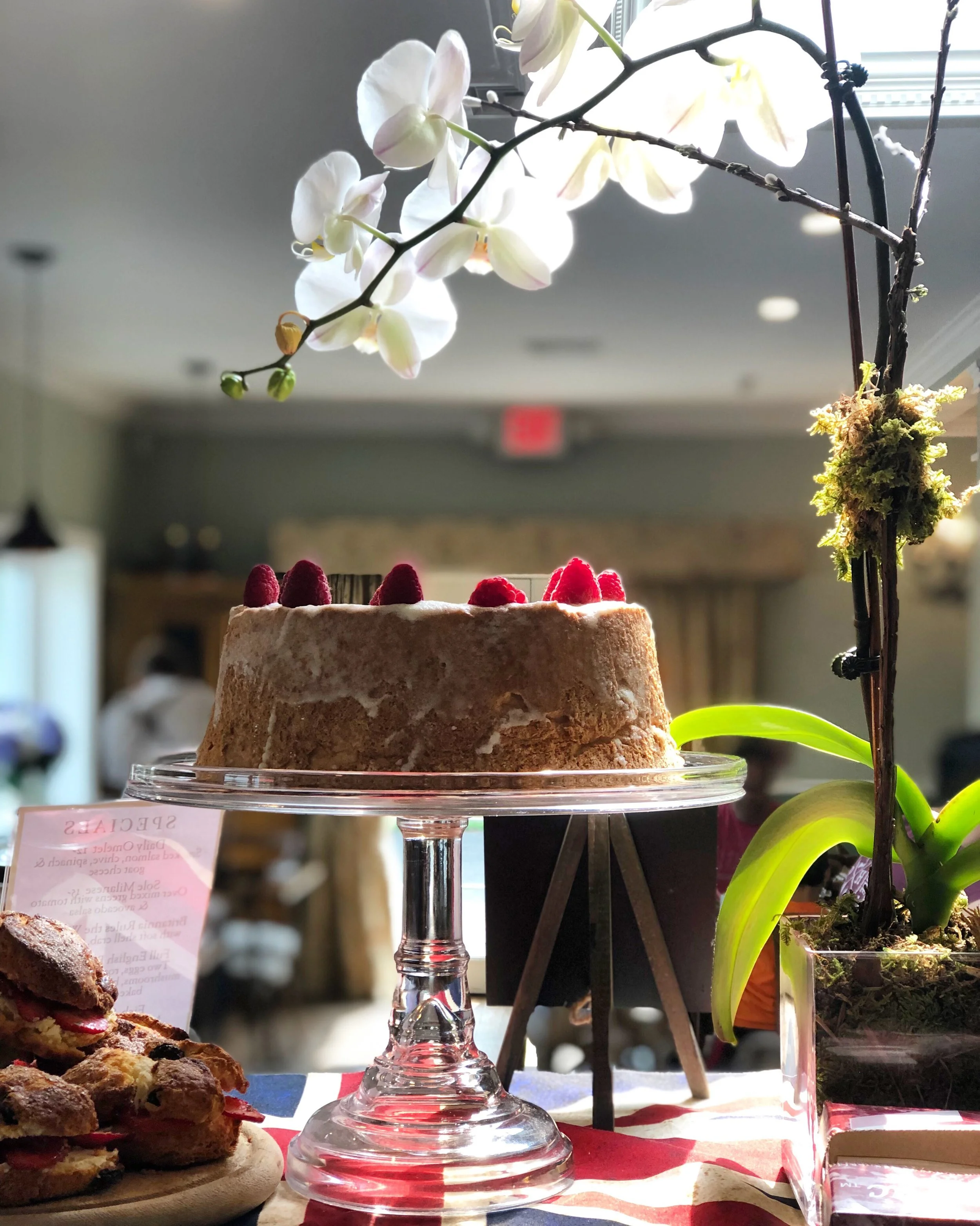 A chocolate cake with raspberries on top, displayed on a glass cake stand, surrounded by orchids and small pastries, with a blurred indoor background.