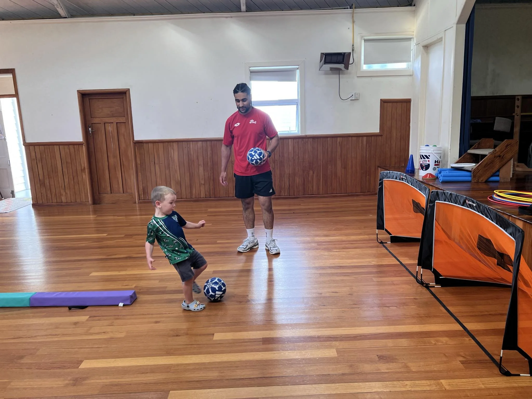A young boy in a soccer jersey kicking a soccer ball while an adult coach watches inside a gymnasium with wooden floors and walls.