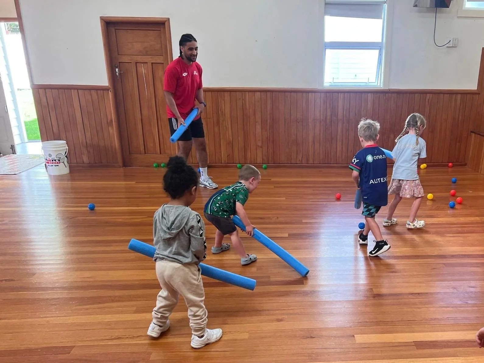 Children playing floor hockey with foam sticks and balls, supervised by an adult in a room with wooden floors and paneling.
