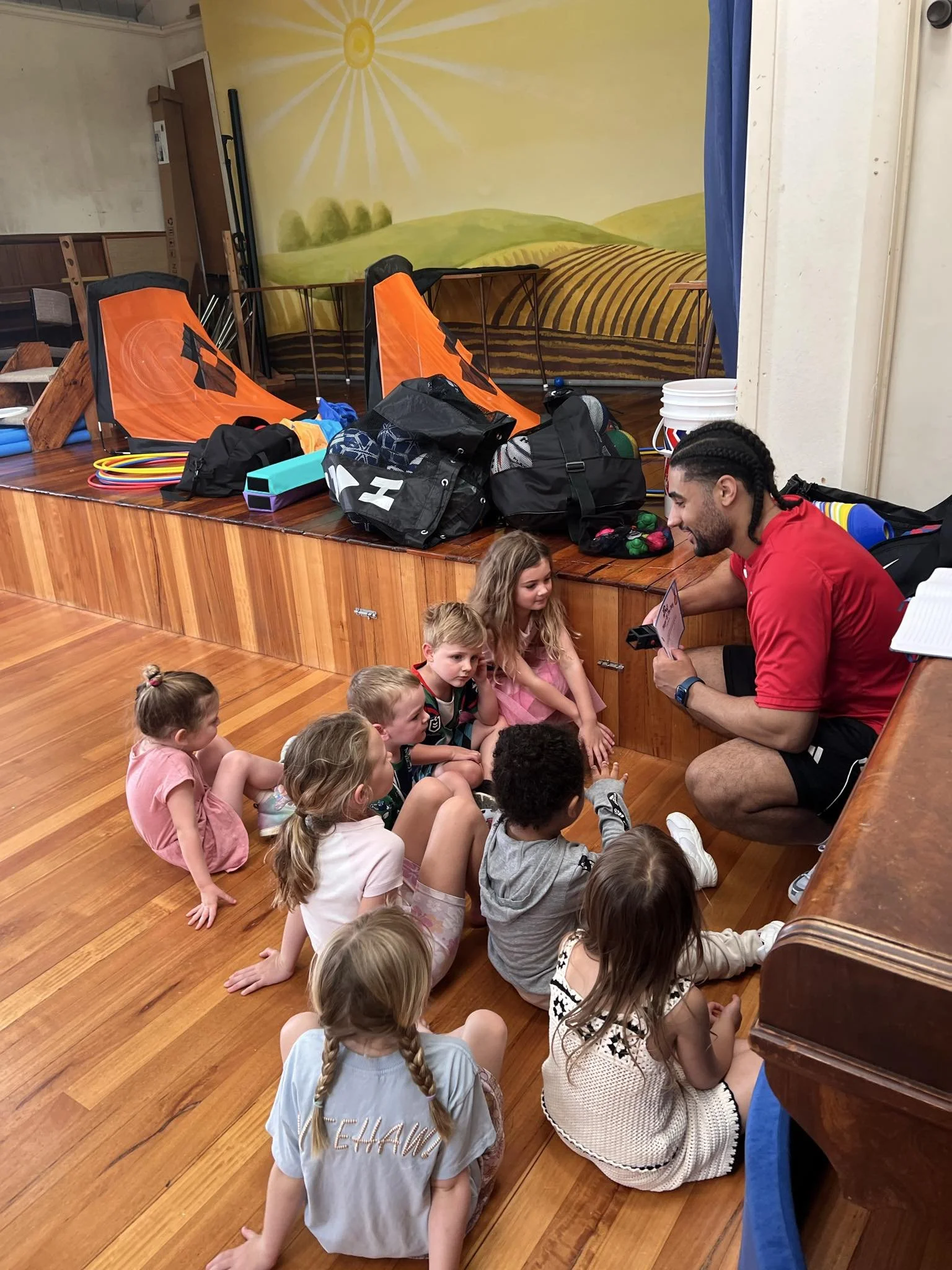 A man with braided hair, wearing a red shirt, is sitting on the floor and reading a book to a group of young children sitting cross-legged on the wooden floor in a room with a mural of a sunny landscape on the wall.