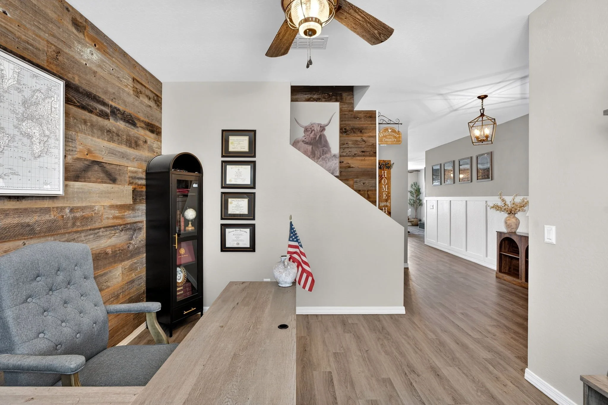 Interior of a home office or dining area featuring wood-paneled walls with framed diplomas, a grey tufted armchair, and a wooden table with a decorative vase and small American flag. In the background, there is a hallway with hanging light fixtures, 