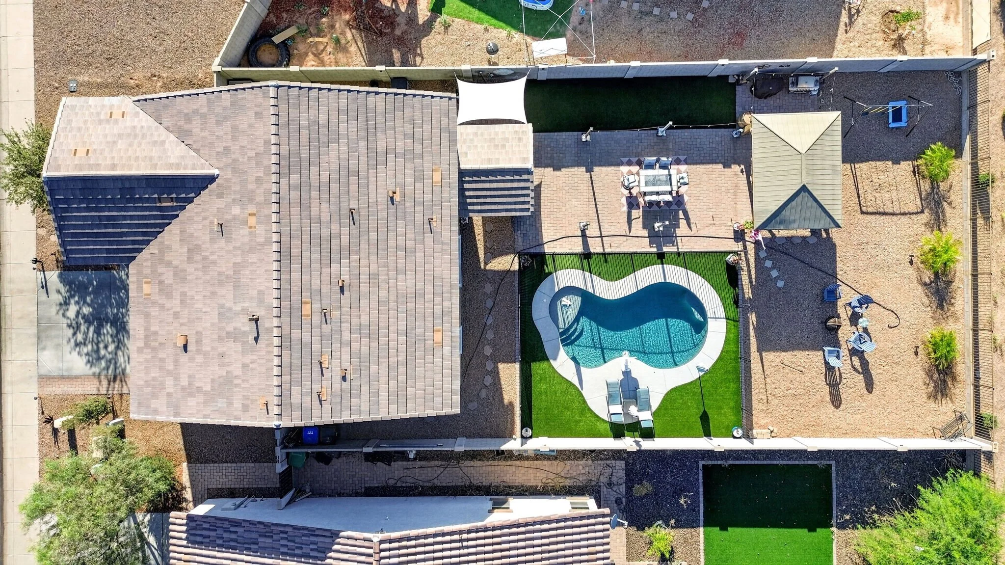 Aerial view of a backyard with a swimming pool, a patio with outdoor furniture, and a shaded area. The yard is enclosed by a fence and contains trees and outdoor equipment.