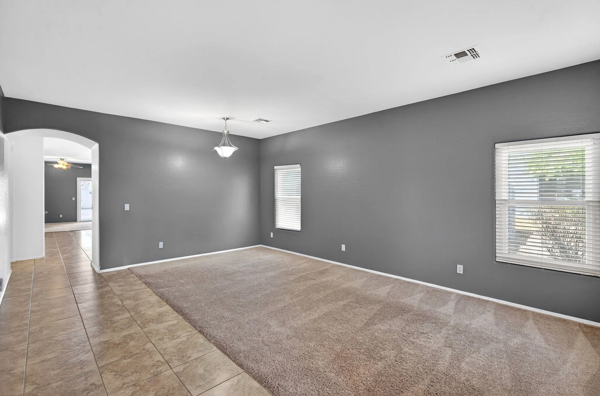 Interior of a room with gray walls, beige carpet, and large windows with white blinds; open doorway leads to another room with tiled flooring and a ceiling fan.