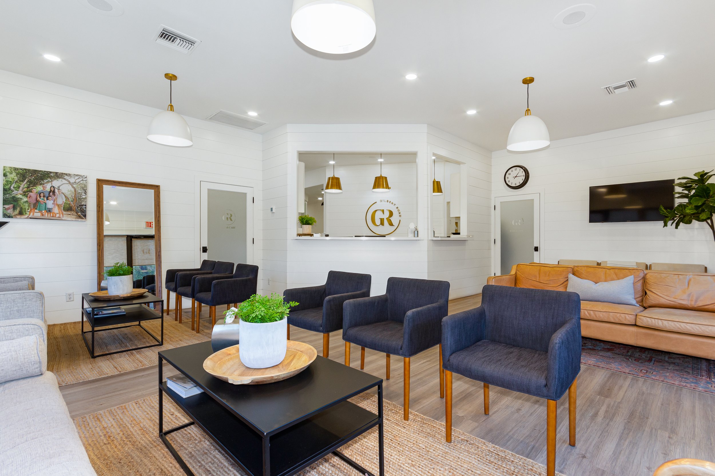 A modern waiting room with black and beige chairs, a black coffee table with a white potted plant, a wooden tray, and magazines, white walls, framed family photo, wall clock, TV, and decorative plants.