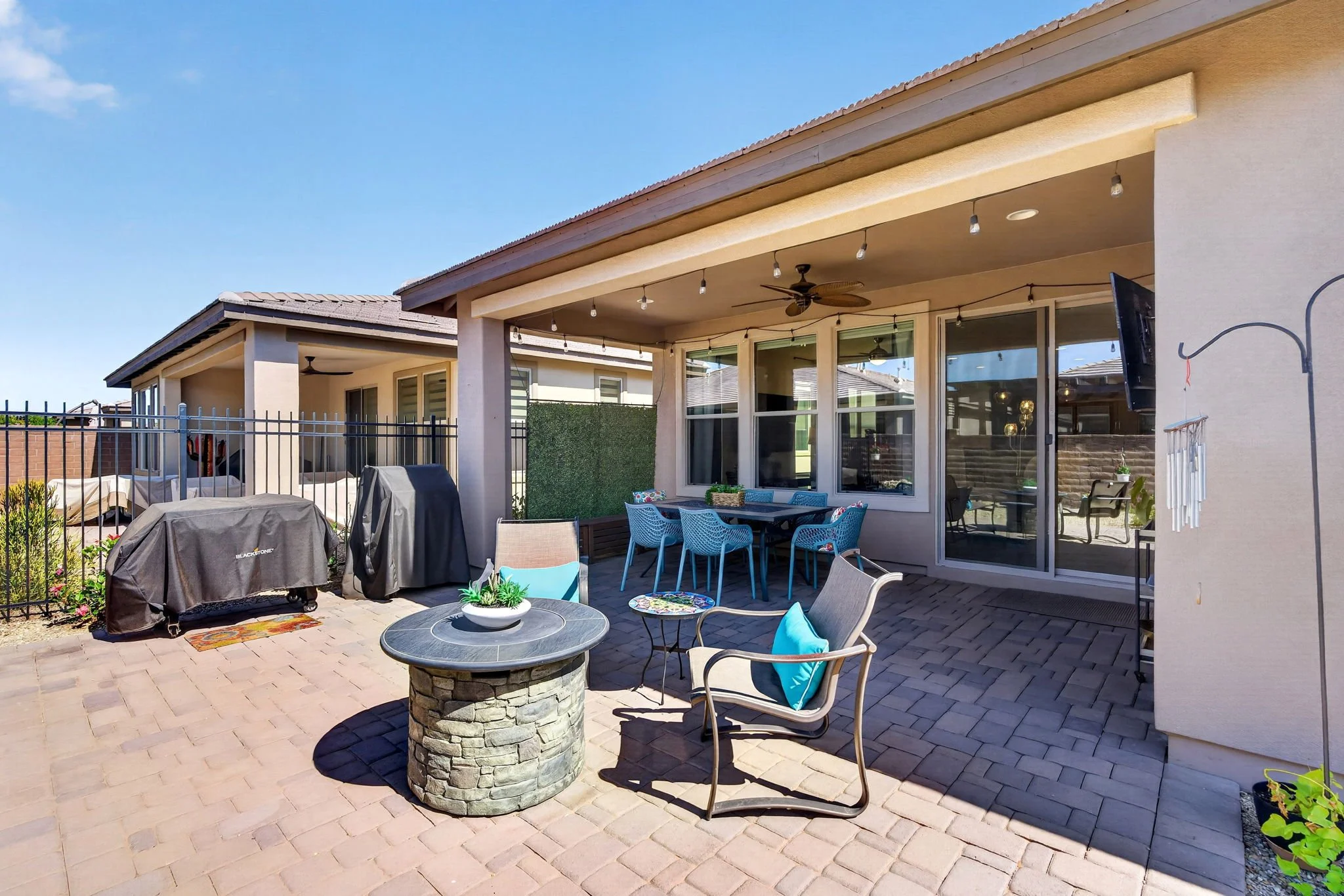 Backyard patio with outdoor furniture, string lights, ceiling fans, grills, and sliding glass doors under a blue sky.