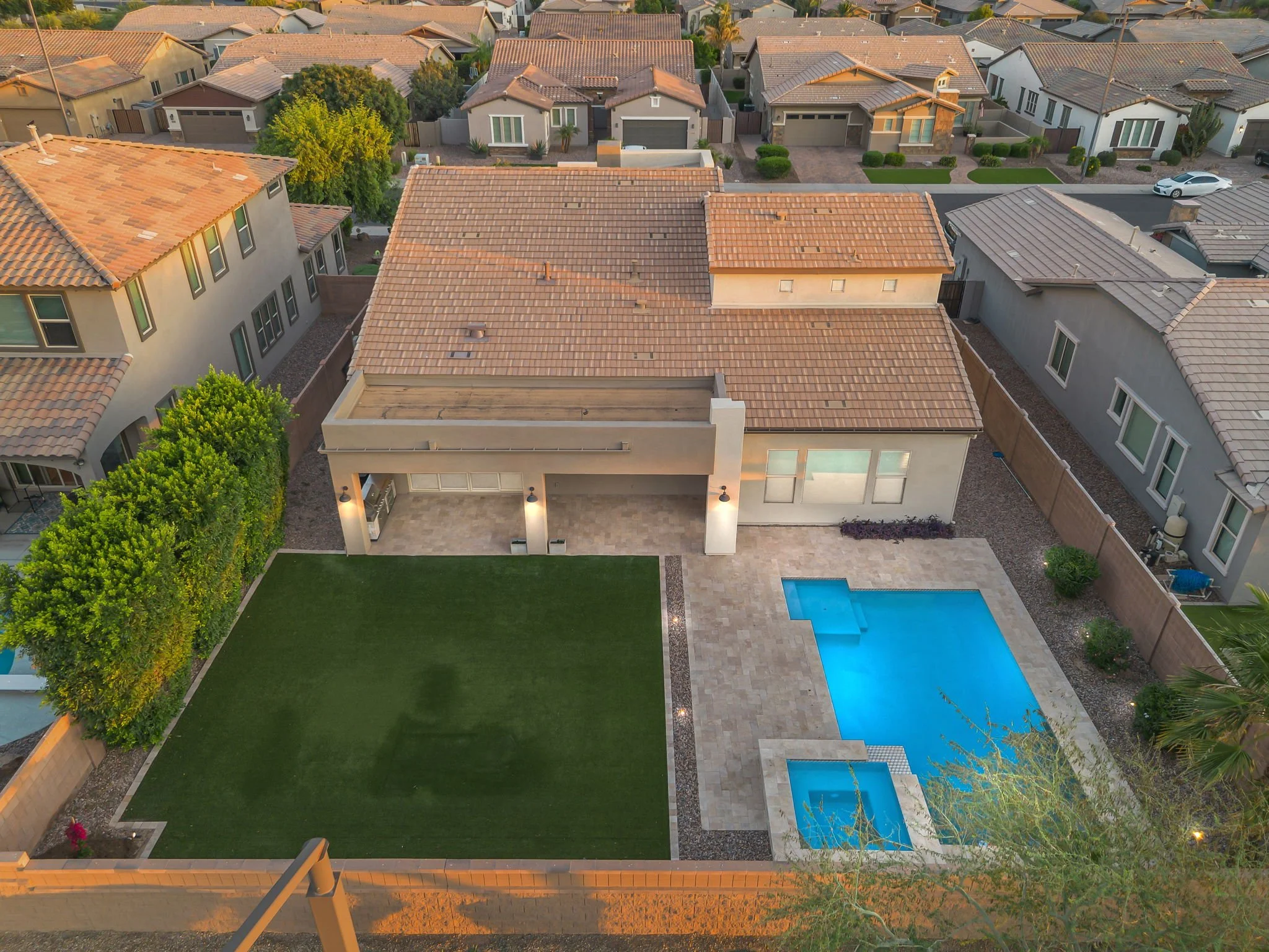 An aerial view of a modern residential backyard with a swimming pool, poolside patio, and a well-maintained lawn surrounded by houses with tiled roofs and landscaped yards.