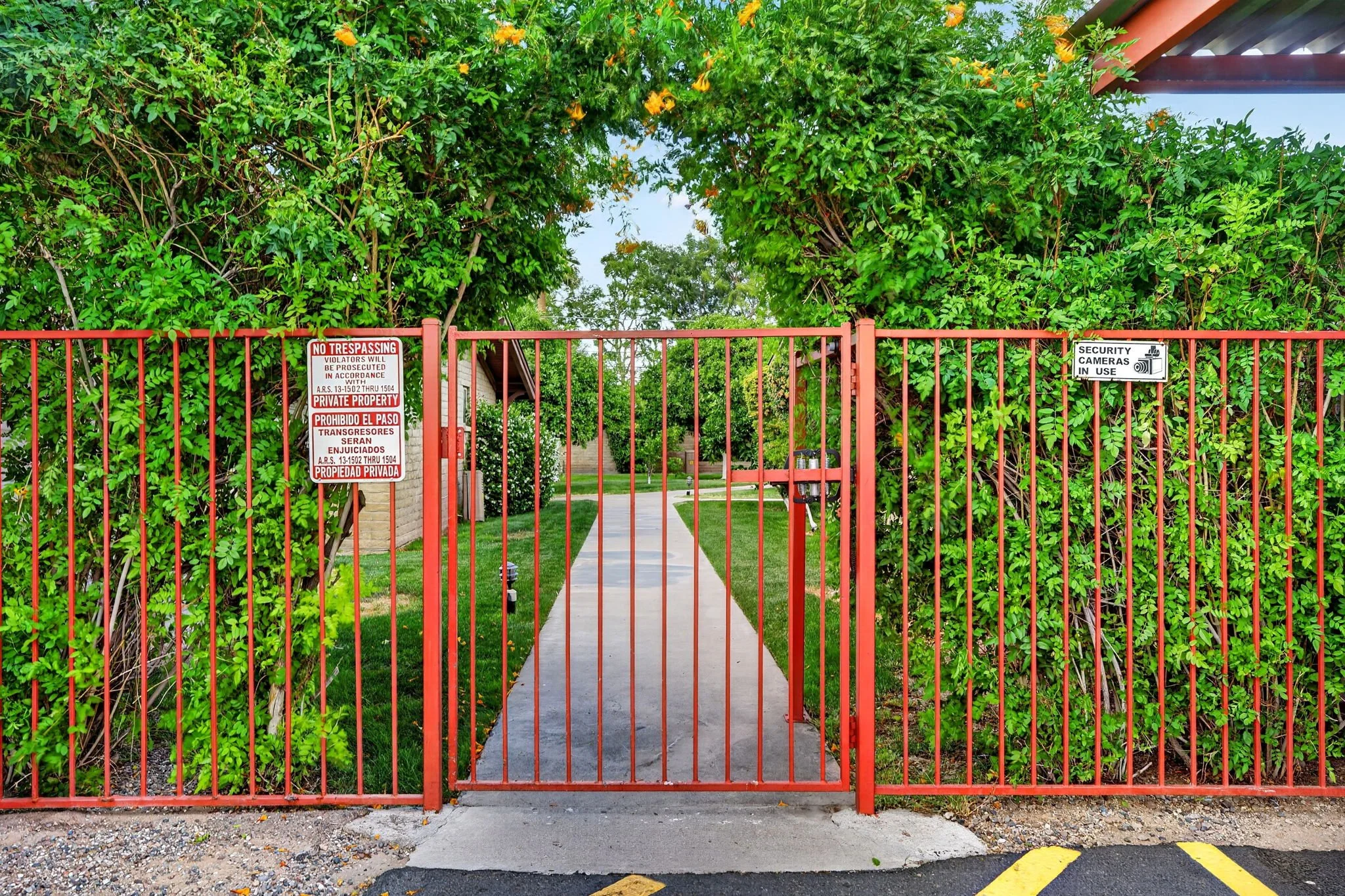 Red metal gate with security camera sign, surrounded by dense green foliage, leading to a sidewalk and entrance of a building.