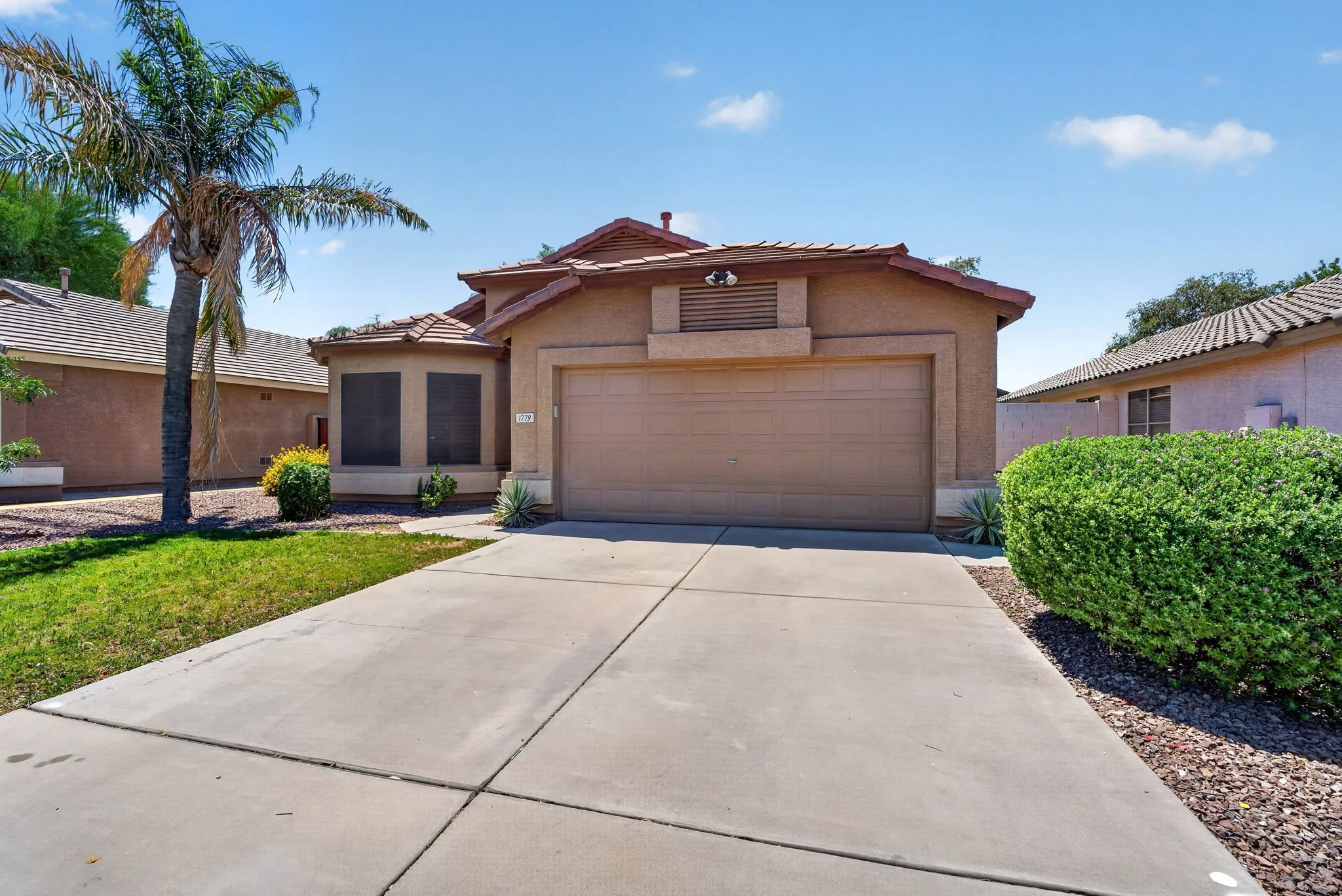 Front view of a beige house with a two-car garage, palm trees, green bushes, and a concrete driveway with a clear blue sky.
