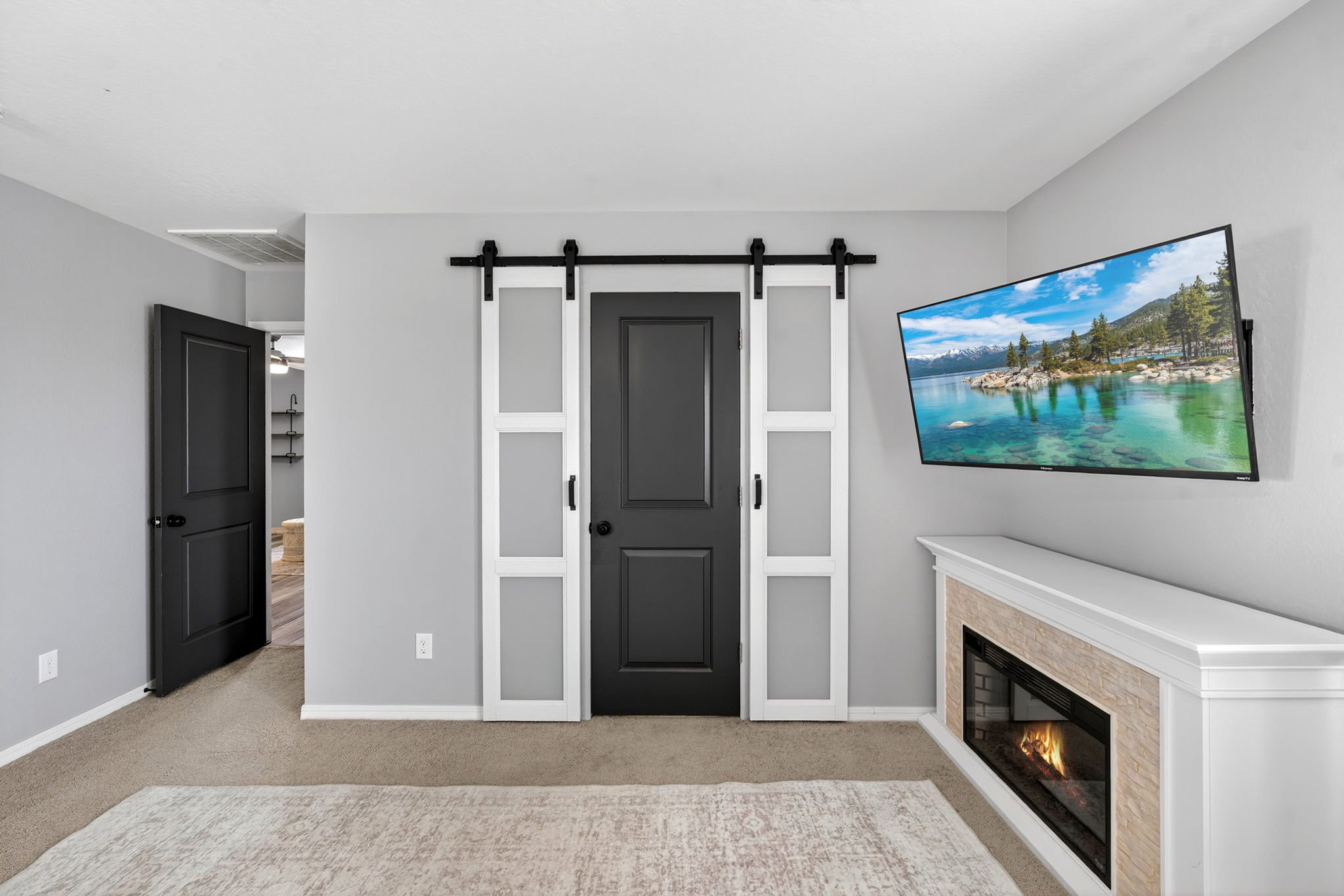 A living room corner with a flat-screen TV showing a scenic mountain lake, a modern fireplace, and a black sliding barn door on a wall painted light gray.