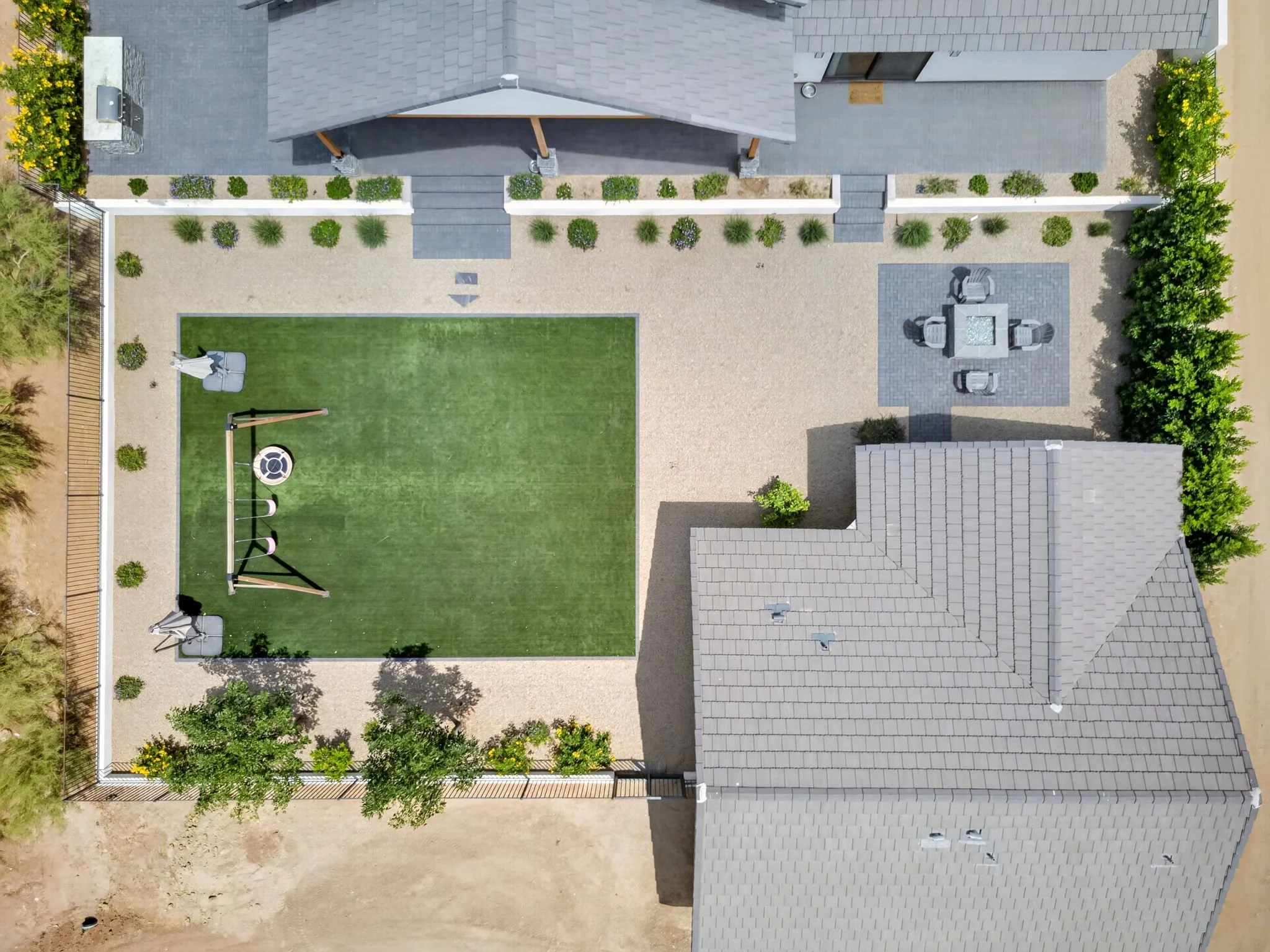 An aerial view of a backyard with a lawn, gravel areas, a patio with outdoor furniture, and a house with gray shingle roofing.