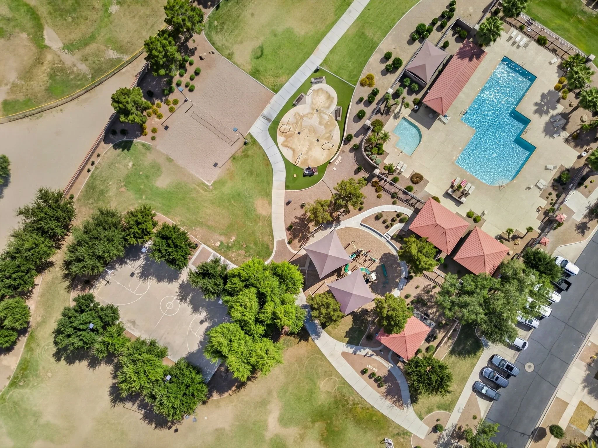 An aerial view of a neighborhood park with a swimming pool, basketball courts, green lawns, trees, and red-roofed structures.