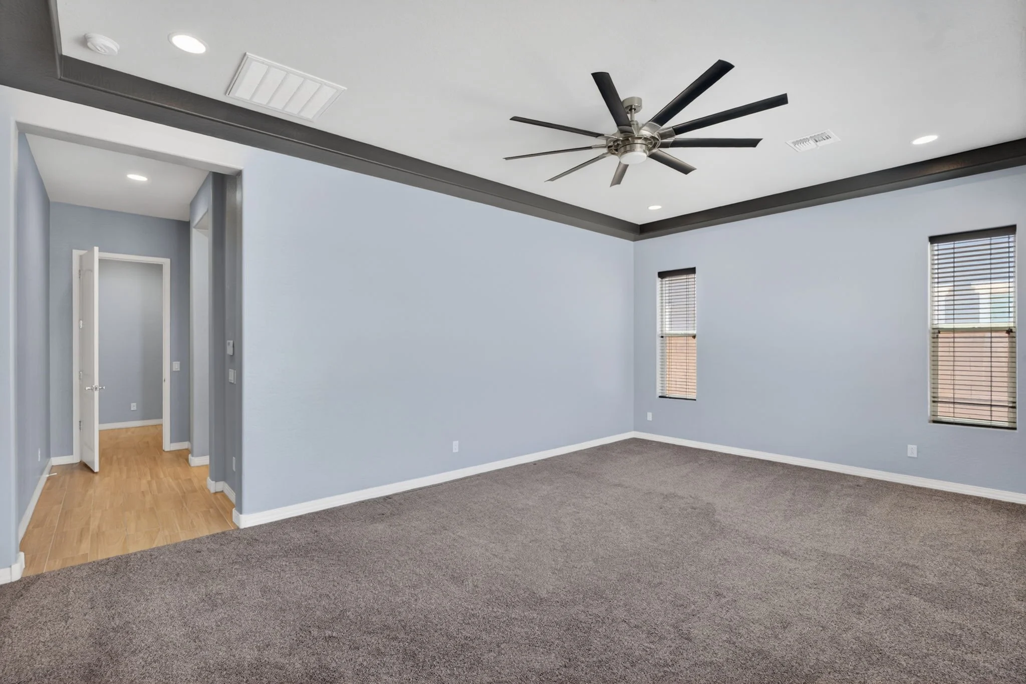 Empty interior room with light blue walls, gray carpeting, and multiple windows with blinds. Ceiling has a modern fan and recessed lighting.