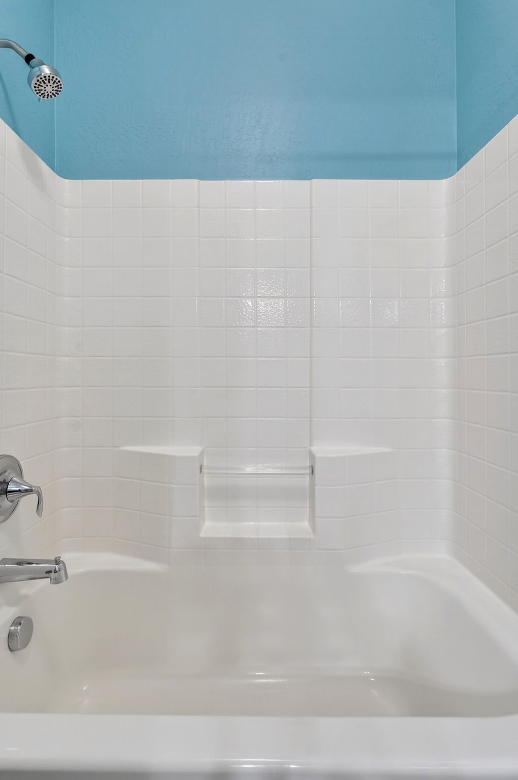 Empty bathtub with white tiled walls and a blue ceiling, featuring a showerhead on the upper left, a faucet and control on the lower left, and built-in shelves in the wall.