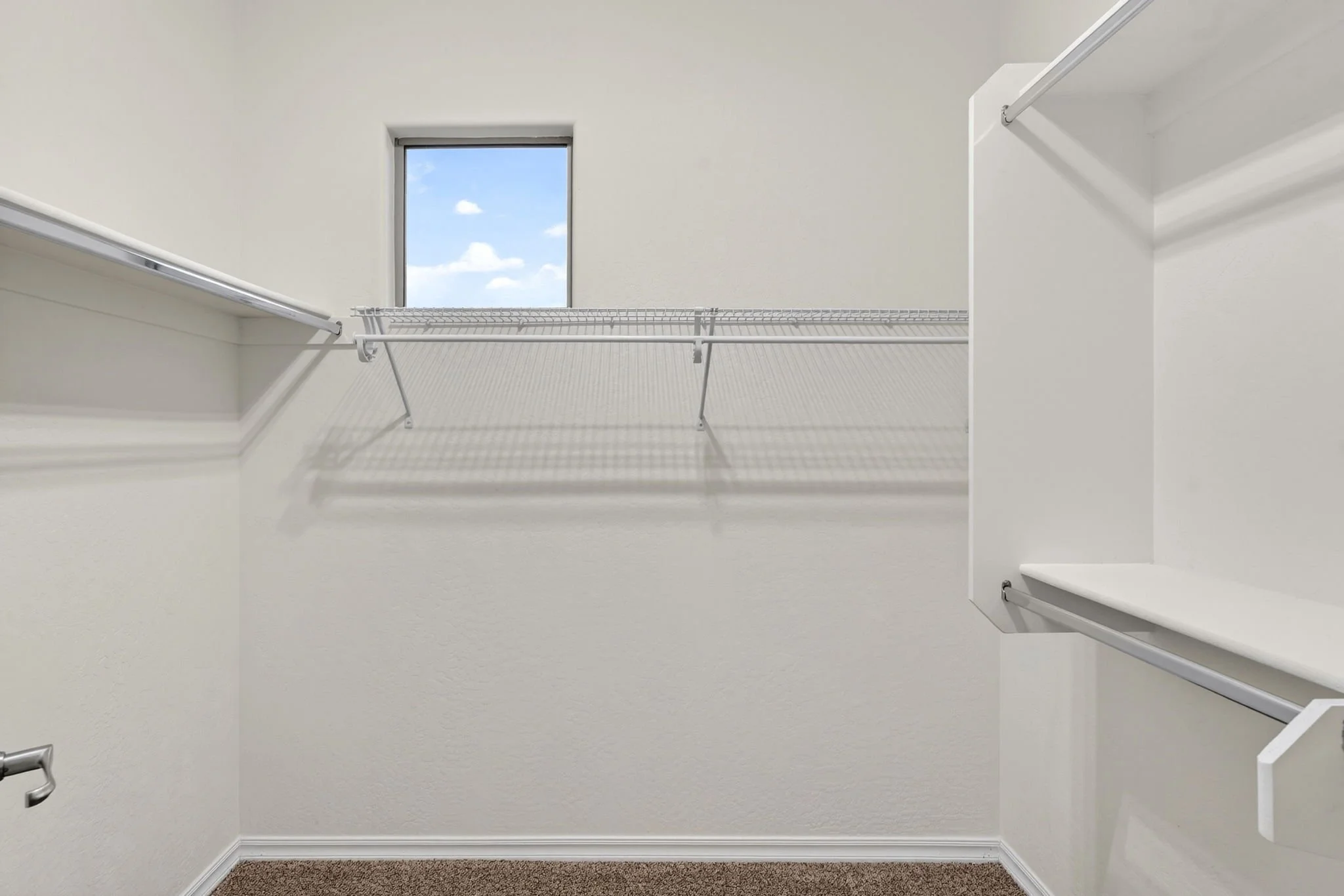 Empty walk-in closet with wire shelving, a small window showing blue sky and clouds, and carpeted floor.