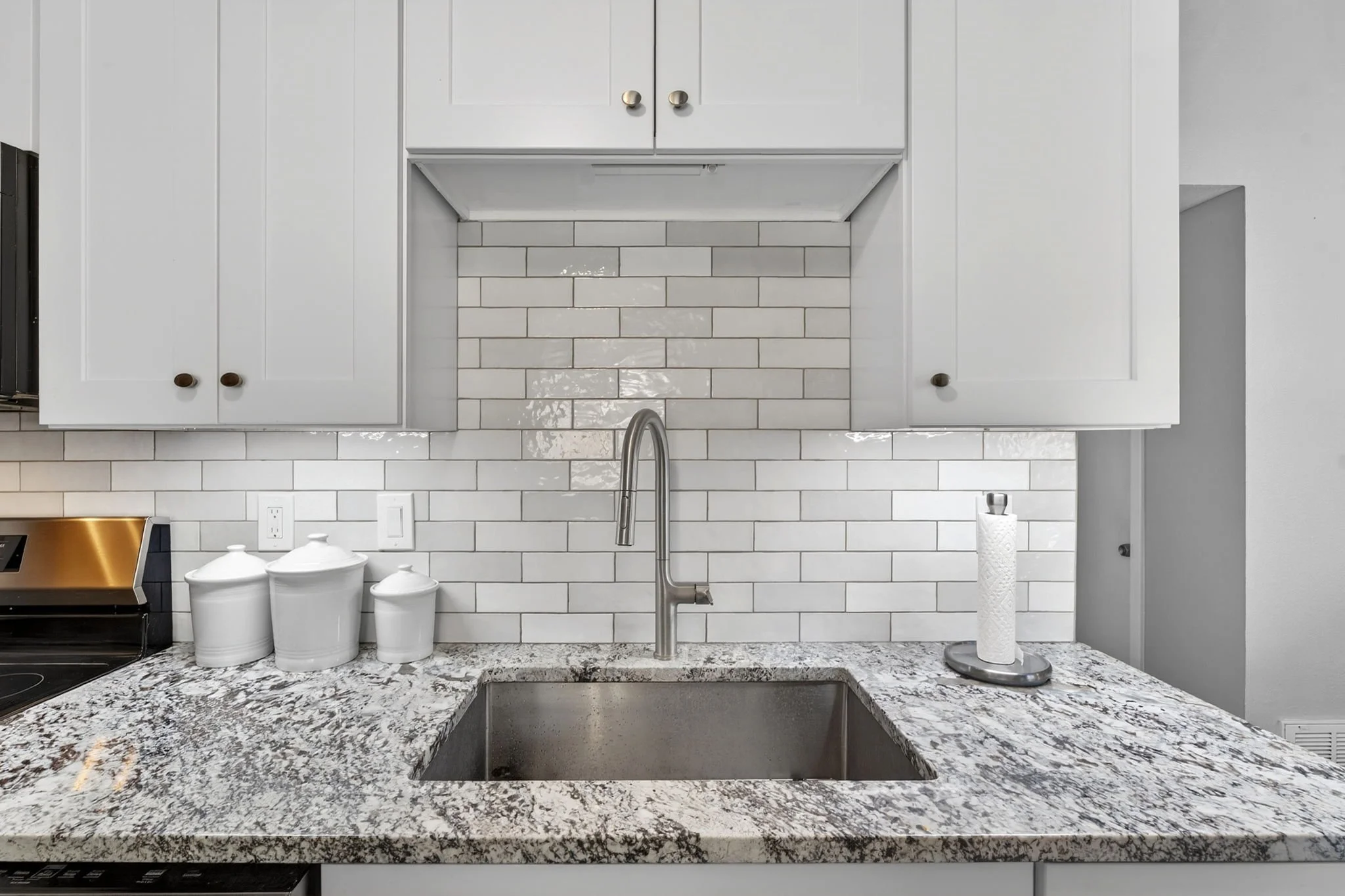 A modern kitchen countertop made of granite with a built-in sink, a stainless steel faucet, a paper towel holder, and white ceramic canisters. White cabinets with round knobs are mounted on a white subway tile backsplash.