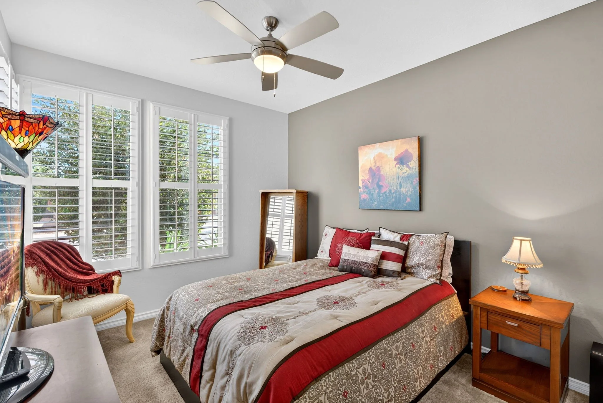 A bedroom with large windows, a bed with a red and beige quilt, pillows, a wooden nightstand with a lamp, a mirror, a chair with a red throw, and a ceiling fan.