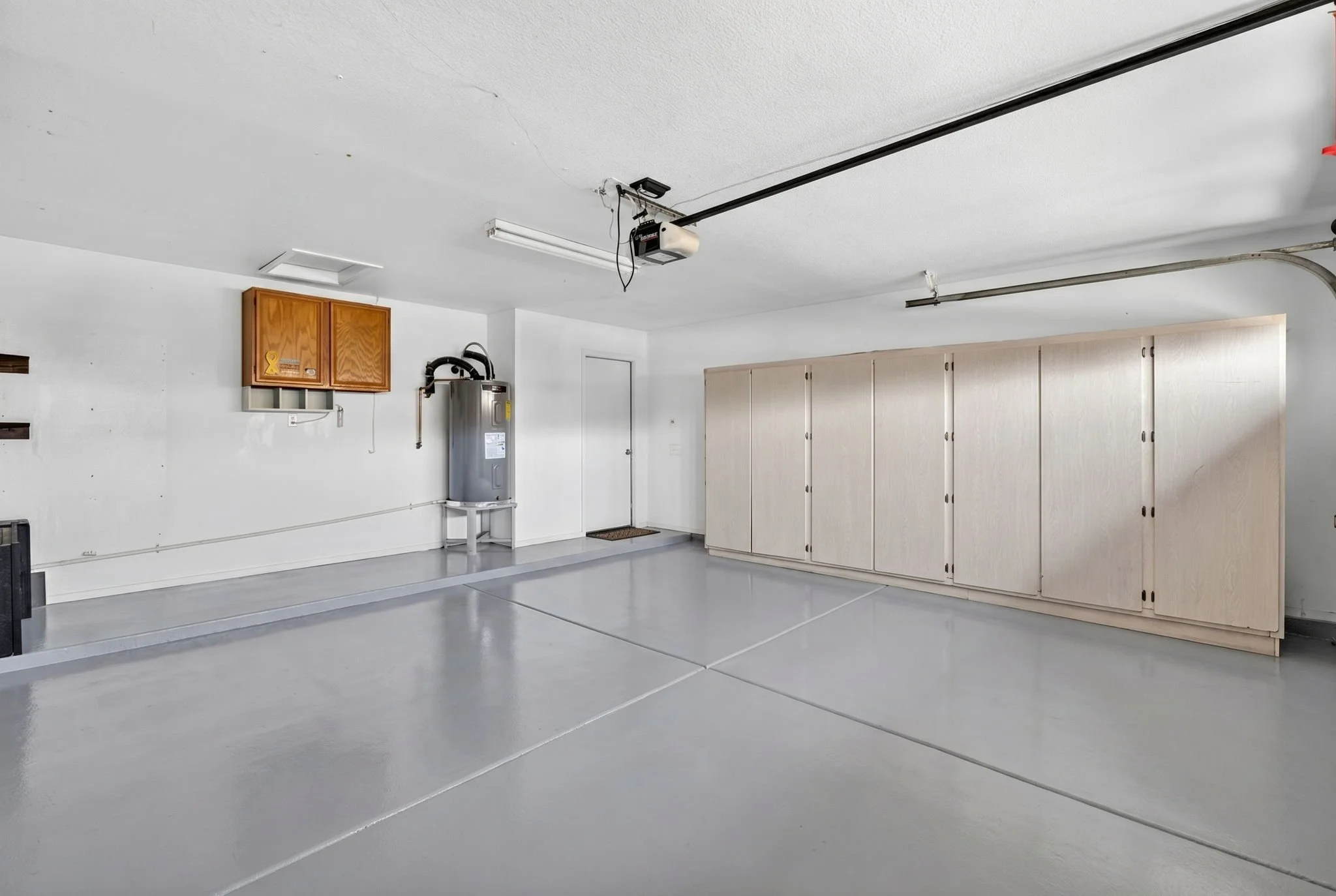 Empty garage with light gray floor, white walls, wooden cabinet, water heater, and large beige storage cabinets.