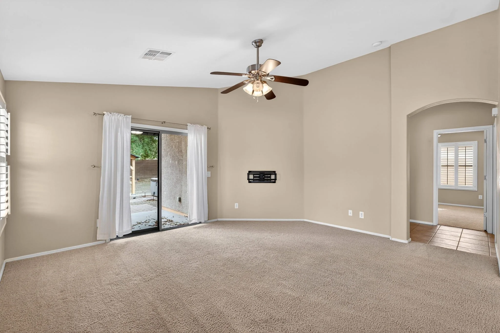 Living room with beige walls, ceiling fan, white curtains, carpeted floor, and sliding glass door leading outside.
