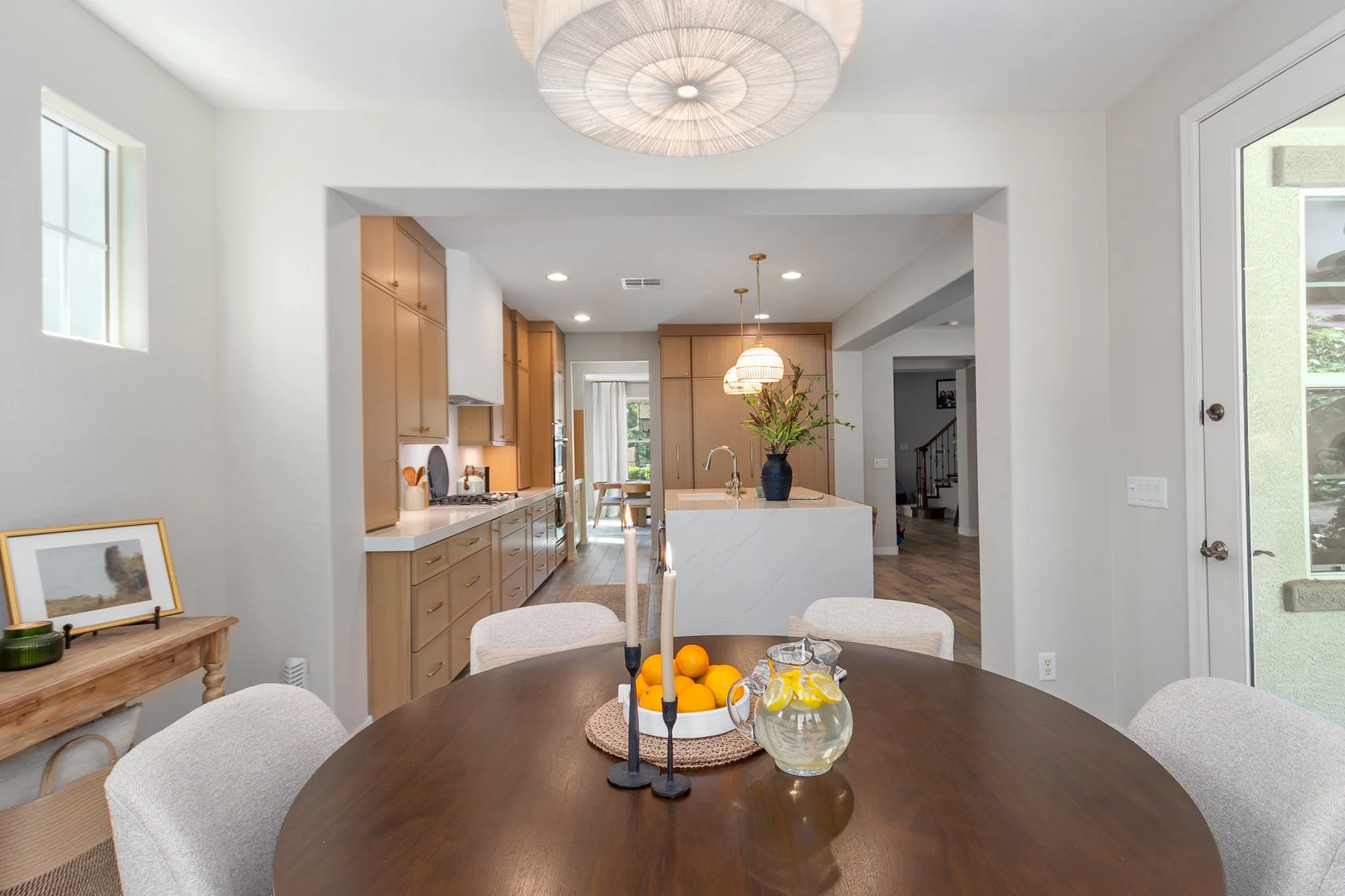 Dining area with a dark wood table, surrounded by light-colored upholstered chairs, with a bowl of oranges and a glass pitcher on the table. The background features a kitchen with light wood cabinets, a white island, and pendant lighting, with a wind
