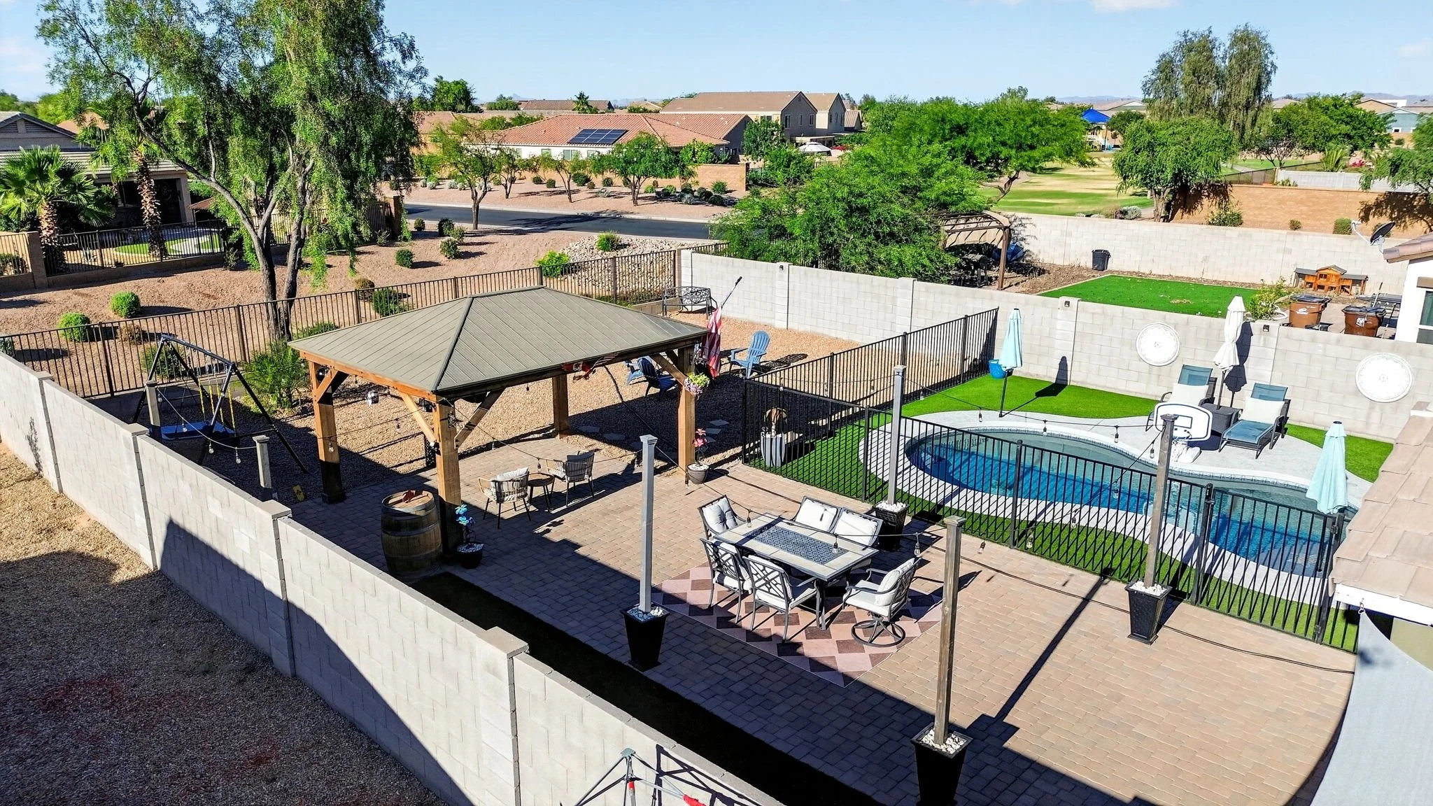 A backyard with a swimming pool, a gazebo, outdoor seating, and various trees and plants, enclosed by a white privacy wall.