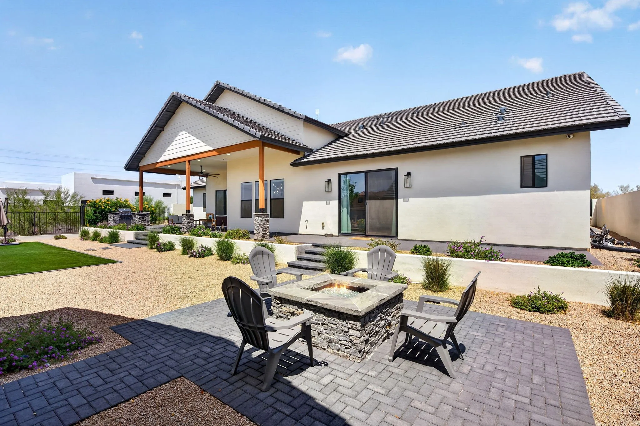 Backyard with a fire pit, four chairs, a manicured lawn, garden, and a modern two-story house with patio and large windows.