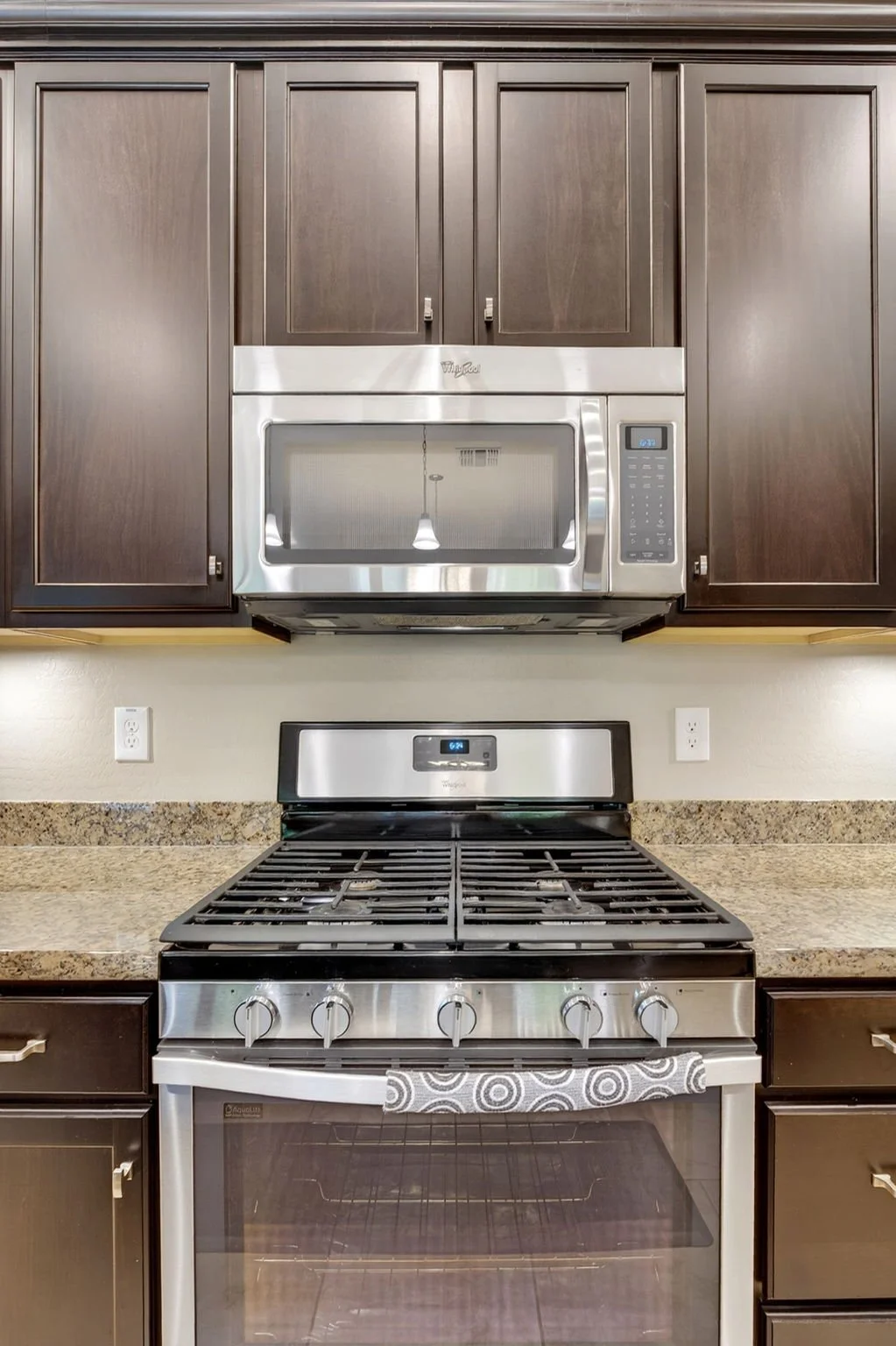Kitchen with dark brown cabinets, granite countertops, a stainless steel microwave above a stove, and two electrical outlets on the wall.