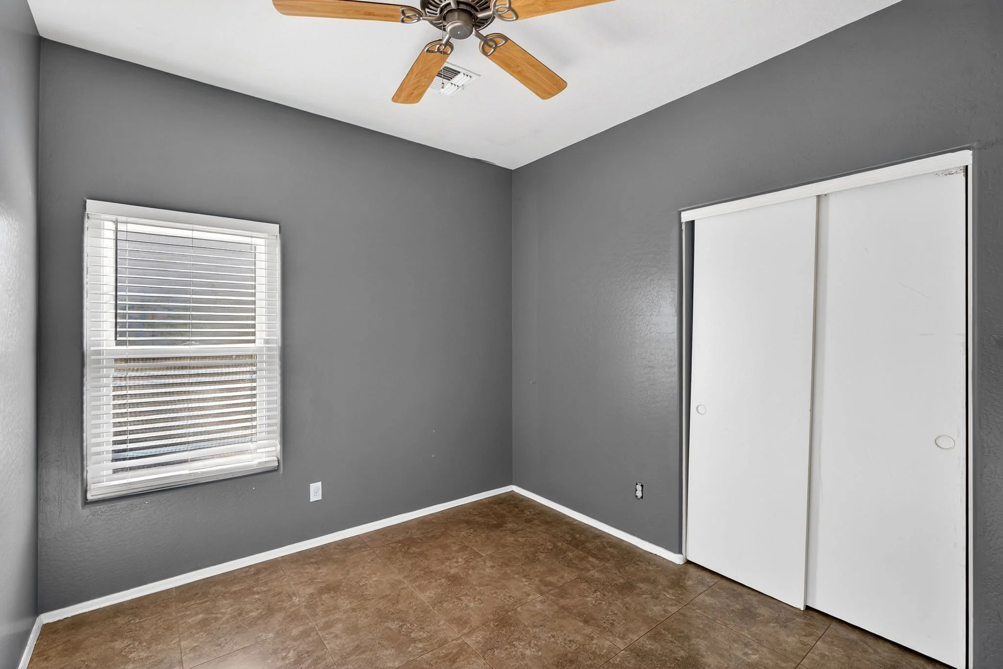 Empty room with a gray wall, white ceiling, two windows with blinds, wood floor, and a closet with white sliding doors. A ceiling fan with wooden blades is mounted on the ceiling.