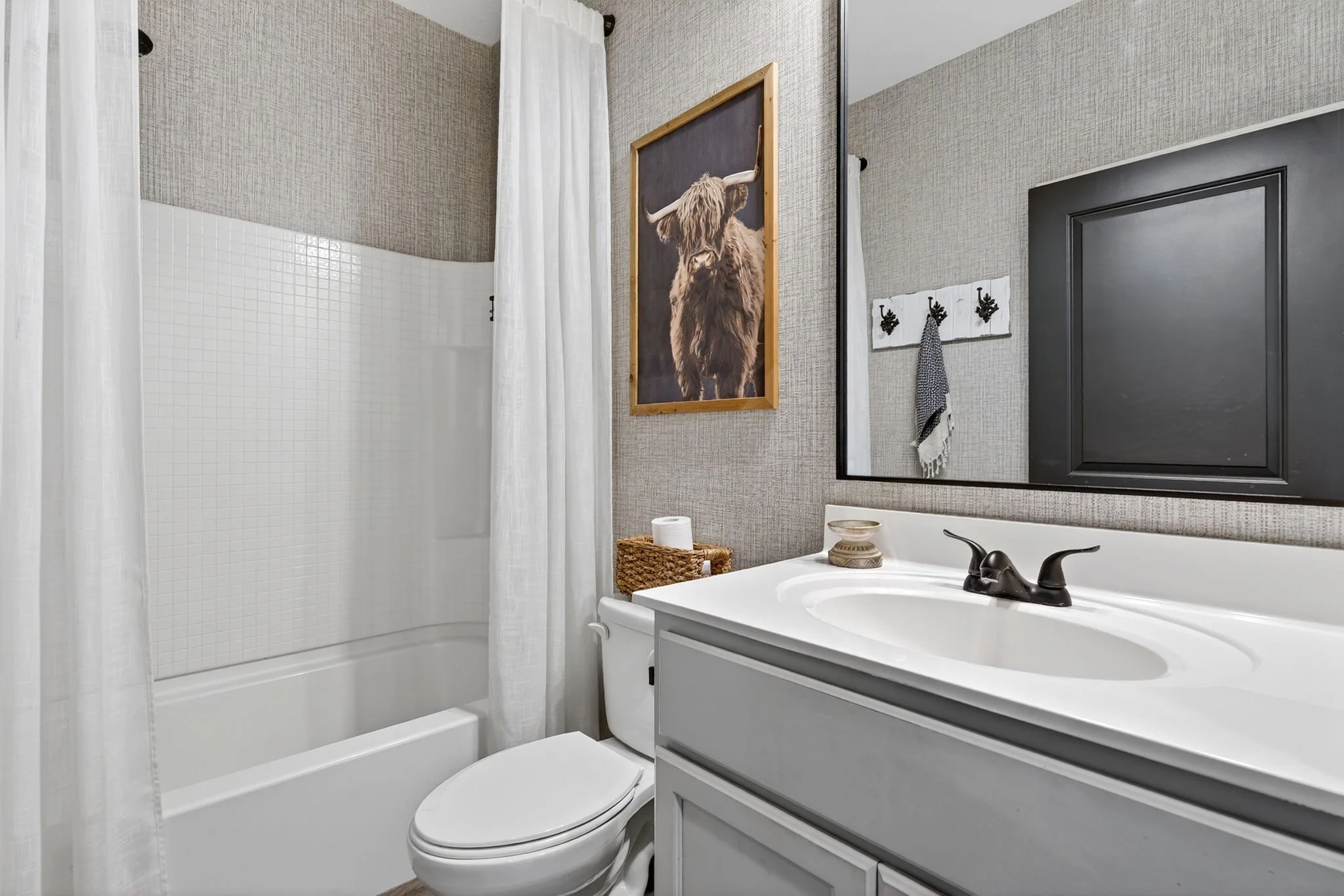 A small bathroom with a white bathtub, shower curtain, toilet, and white vanity with a sink. The walls are textured gray, and there is a framed picture of a highland cow on the wall. A mirror is visible above the sink, and a basket with toilet paper 