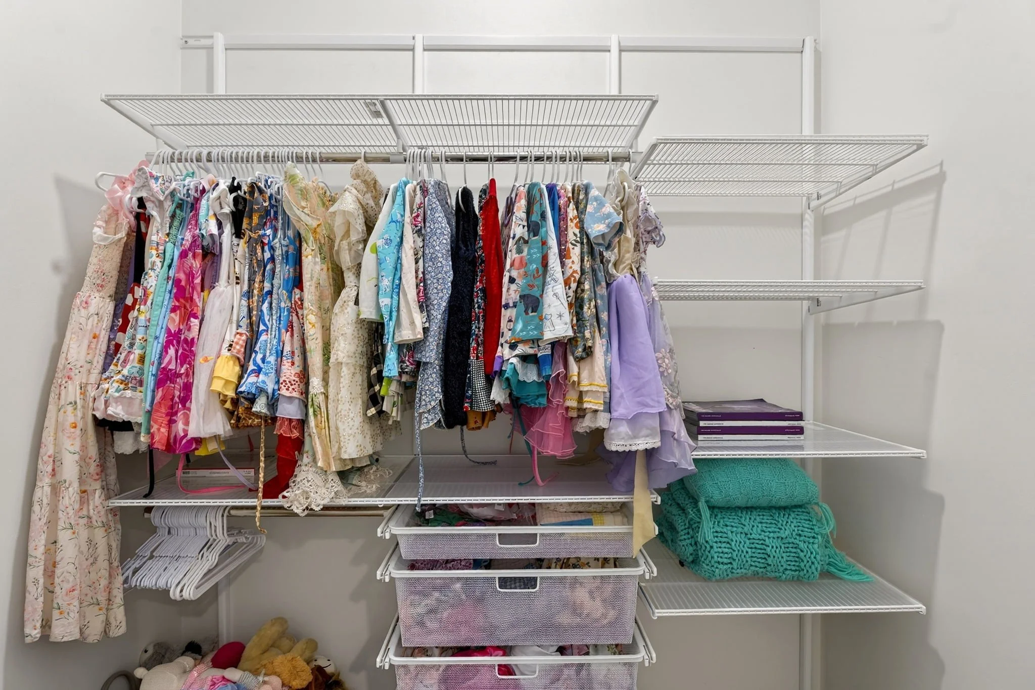 A closet with white wire shelves holding children's clothes in various colors and patterns, with storage bins on the lower shelves and stuffed animals and additional items on the floor.
