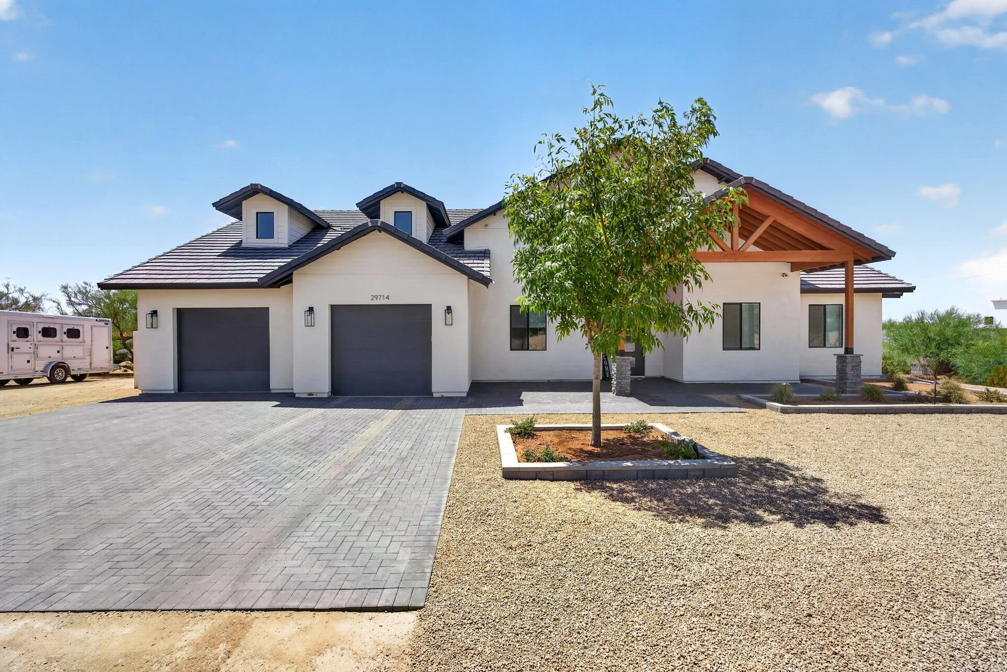 Modern house with a two-car garage, driveway, small front yard with a tree, and a covered porch, under a partly cloudy sky.