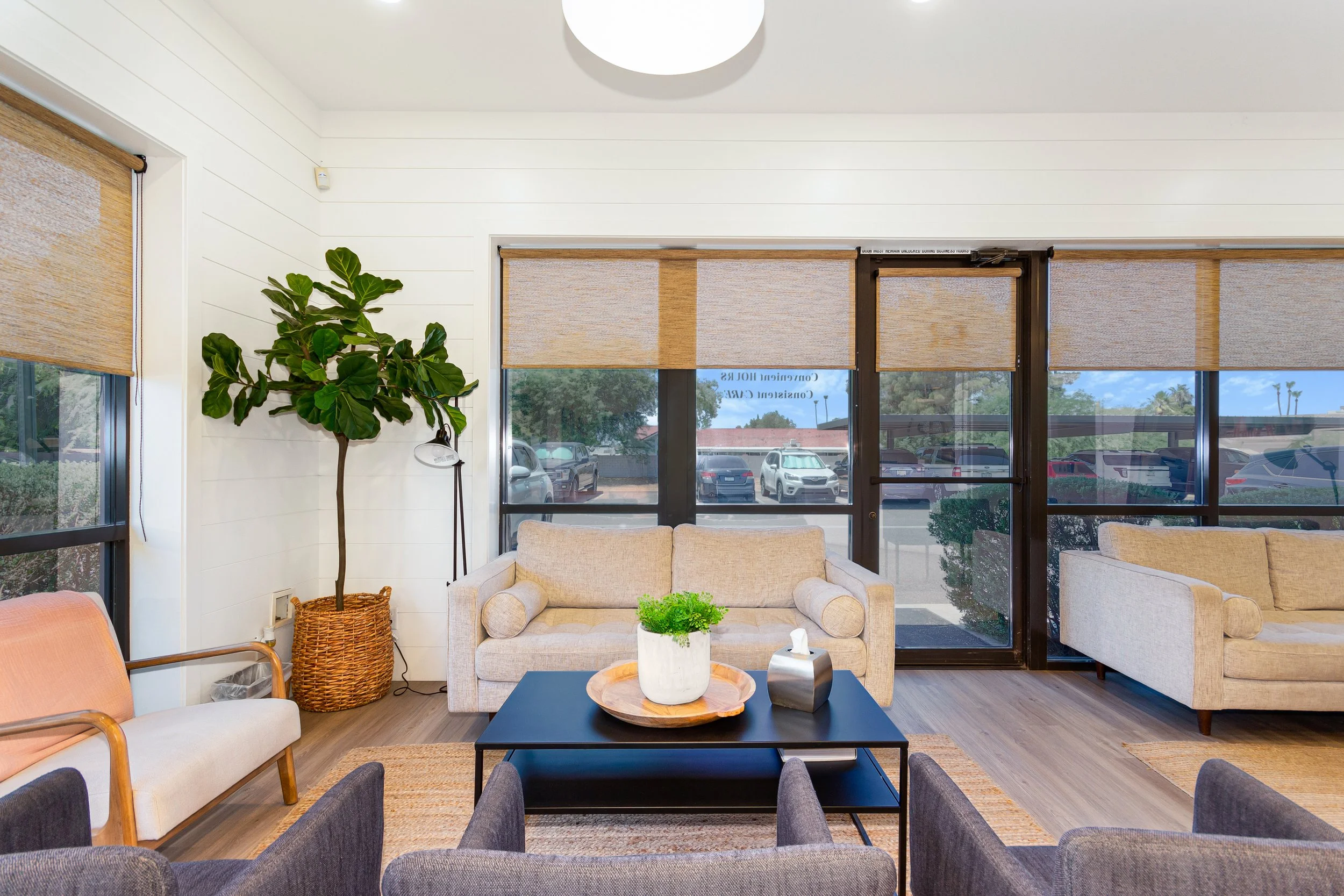 A cozy waiting room with beige sofas, a black coffee table with decorative bowls, a potted plant, and large windows with blinds showing a parking lot outside.