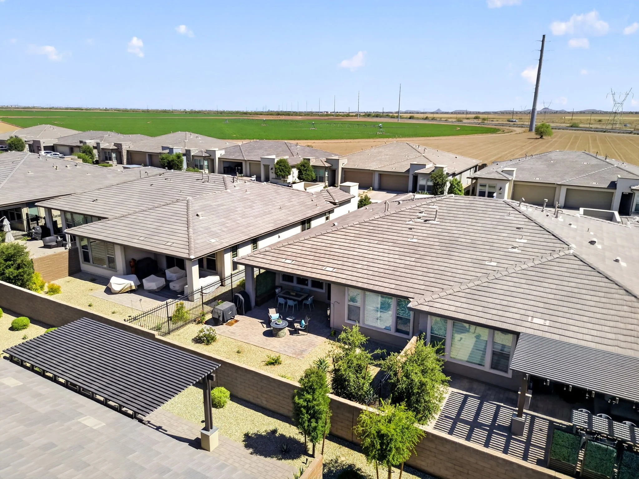 A residential neighborhood with single-story houses featuring beige exteriors and gray tiled roofs. The backyards have patios, outdoor furniture, and small trees. Beyond the houses, there are fields and flat land with power lines and a clear blue sky