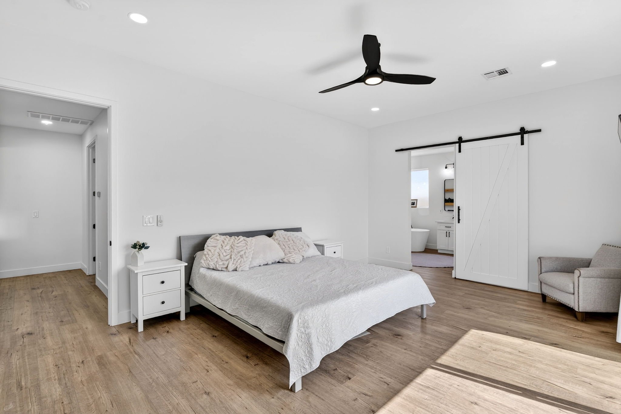A modern bedroom with white walls, a bed with white bedding and throw pillows, nightstands, a ceiling fan, and a barn door leading to a bathroom.